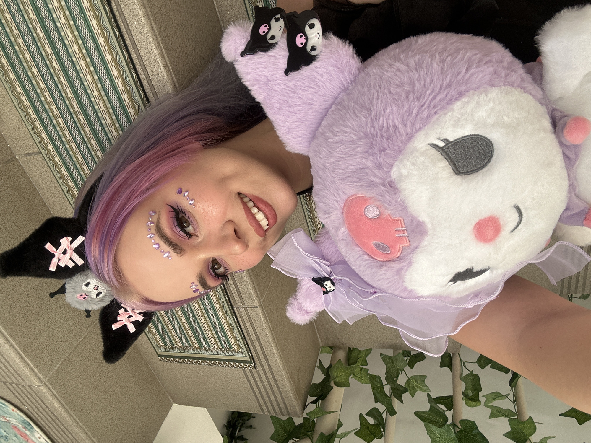 A young woman with lavender-pink hair is holding a purple and white plush toy, possibly Kuromi. Her face is angled towards the camera, with glitter makeup around her eyes. She wears a black headband with Kuromi-themed embellishments. The background includes patterned tiles, possibly a rug, and a wall decorated with artificial ivy. It's not possible to pinpoint the specific location in Zagreb, Croatia, from this image alone.
