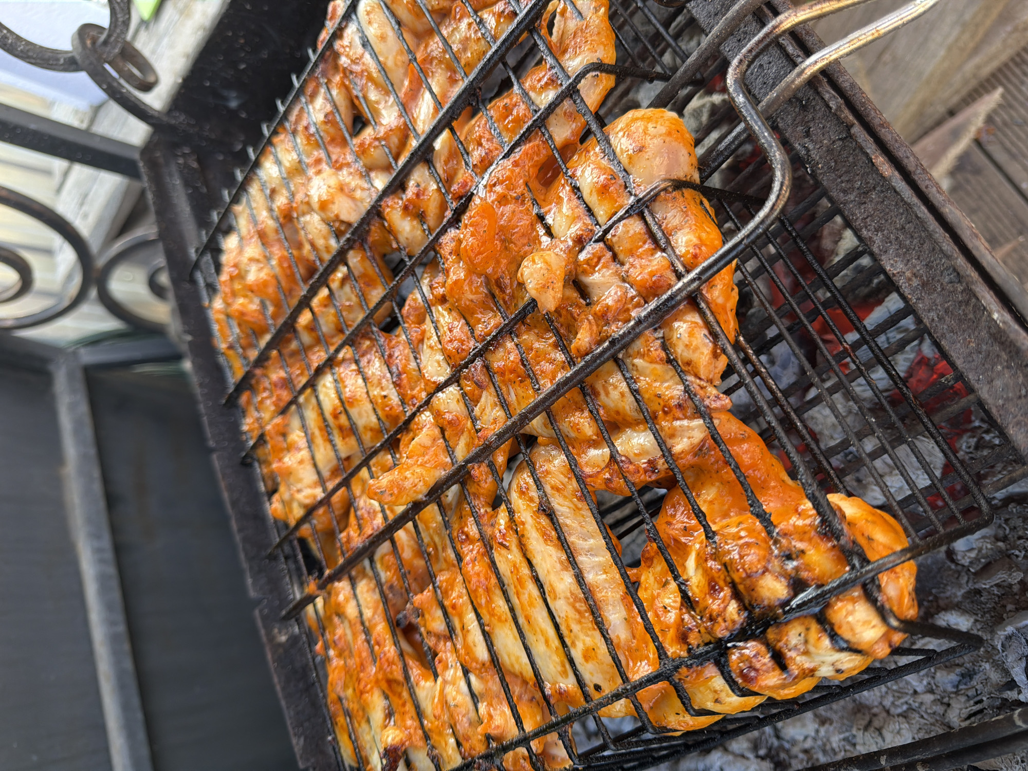 A grill filled with rows of seasoned, cooked chicken pieces sits on a metal grate, likely over hot coals, based on the residue below. The grill is part of an outdoor cooking setup in an urban environment in Chaikovsky, Russia. The grill has ornate metalwork on its side. The scene is brightly lit, suggesting outdoor daylight.
