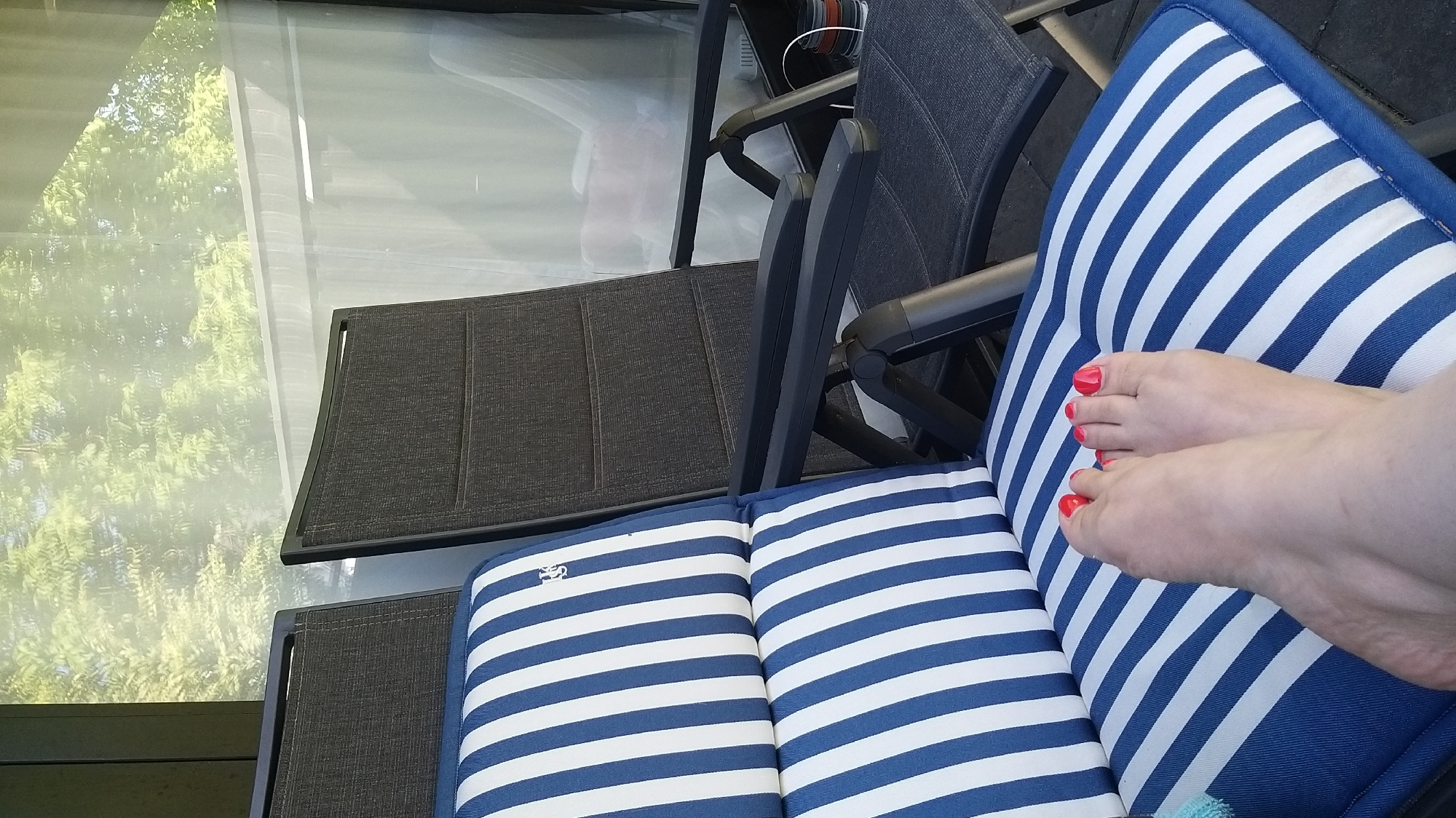 The image shows a close-up of a pair of feet with red nail polish resting on a blue and white striped cushion on a patio chair. Another dark gray patio chair is visible behind it. The background suggests an outdoor setting with trees and a translucent curtain. The image appears to be taken on a sunny day. The location is not identifiable as Krefeld, Germany from the visual information provided.