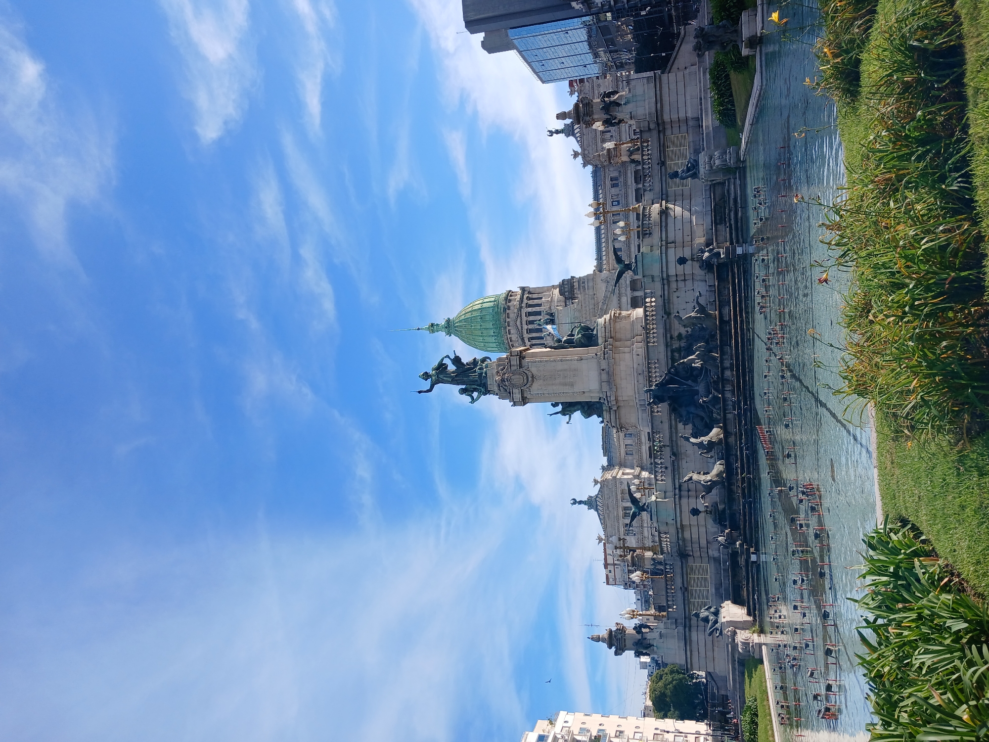 The image shows the Plaza del Congreso in Buenos Aires, Argentina. The Argentine National Congress building is visible with its green dome. In the foreground is a fountain with several water jets. The sky is blue with streaks of clouds. Buildings are visible in the background. Green grass and plants line the edge of the fountain.
