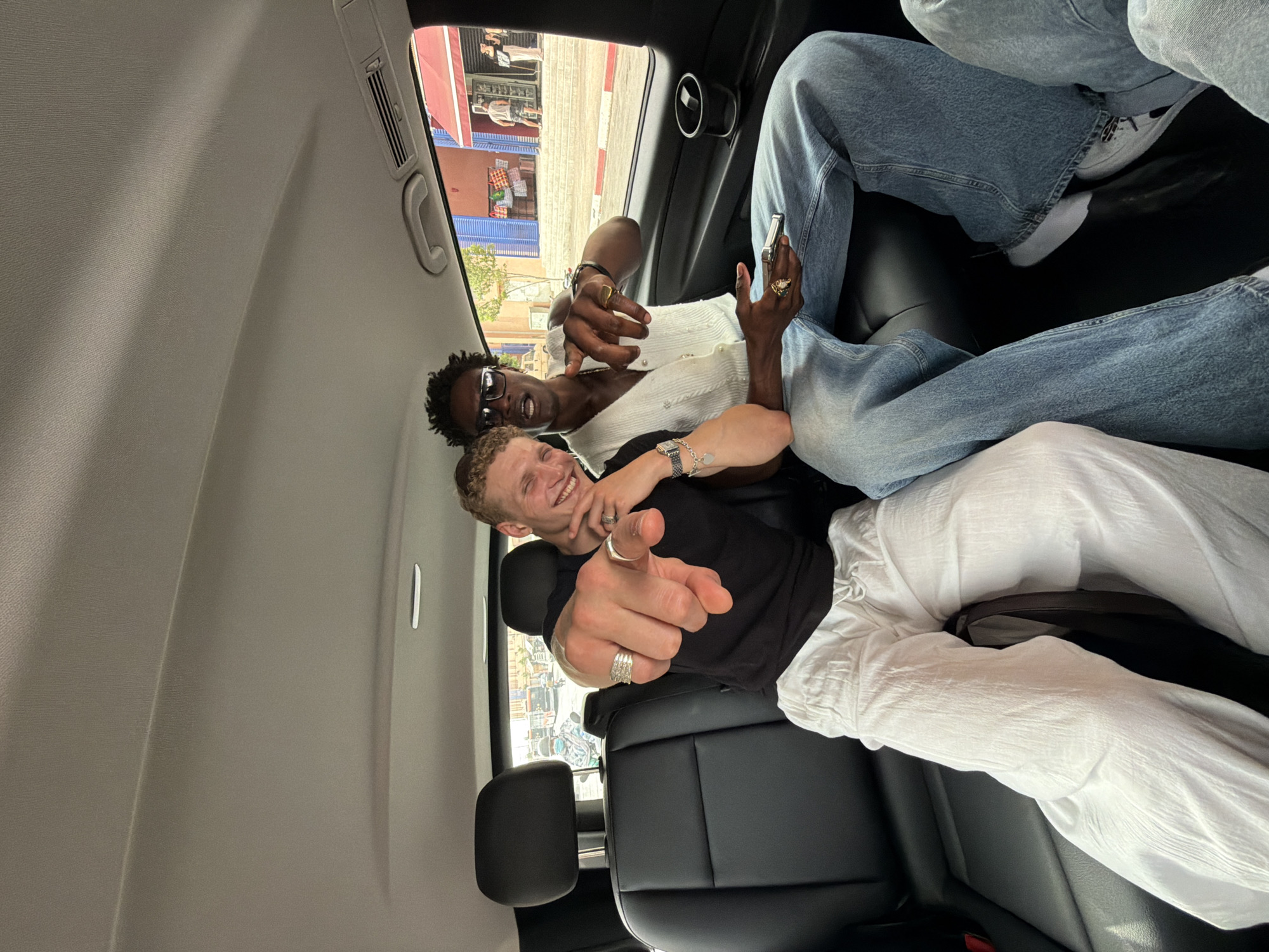 Two young men are seated in the back of a vehicle, presumably a taxi or private car, in Marrakech, Morocco. The man on the left, with fair skin and curly blonde hair, is wearing a black t-shirt and loose white linen pants. He is smiling and pointing directly at the camera with his left hand, which is adorned with several rings. The man on the right, with dark skin and dreadlocks, is wearing sunglasses, a white sleeveless vest over a dark shirt, and light blue jeans. He is also looking at the camera and appears to be speaking. He holds a silver rectangular object, possibly a phone, in his right hand and has rings on his fingers. They are both seated on black leather seats. Through the window of the car, a street scene in Marrakech is visible, featuring traditional Moroccan architecture with blue and red facades, shops with goods displayed, and people walking by. The overall impression is a candid snapshot of friends enjoying their time in the city.