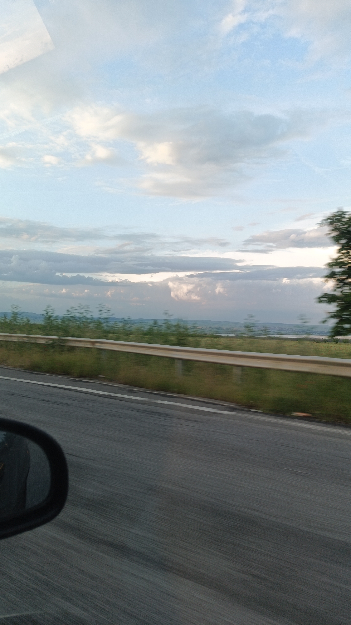 The image, likely taken from a moving vehicle, shows a view of the landscape in Burgas, Bulgaria. The asphalt road takes up a significant portion of the lower half of the frame, with a visible white lane marking. A car's side mirror is partially visible on the left side of the image. A metal guardrail runs parallel to the road, with a grassy area and low-lying vegetation beyond it. In the distance, a faint horizon line is visible beneath a cloudy sky with varying shades of gray and white. The sky occupies the upper half of the image. A blurred tree appears to be on the right.