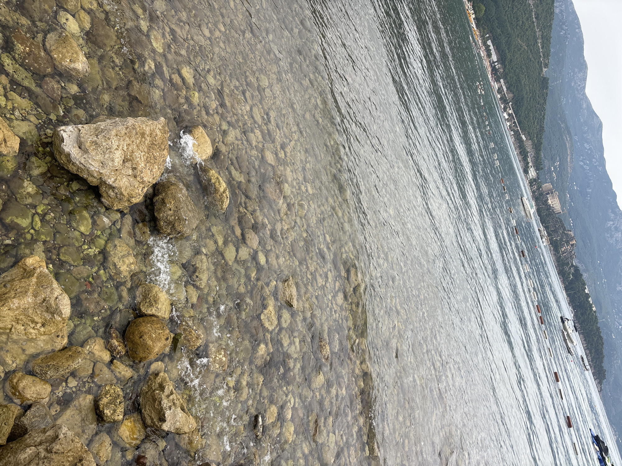 A shallow, rocky beach leads into the sea with the city of Budva, Montenegro visible in the background. The water is clear, revealing a seabed covered in smooth, rounded stones of various sizes and colors. Small waves gently lap the shore, creating white foam around some of the larger rocks in the foreground. In the distance, the coastline is lined with buildings and lush green hills rise to meet a hazy sky. Several buoys mark an area of the sea, and a few small boats are anchored further out. The scene depicts a typical coastal view in the region.