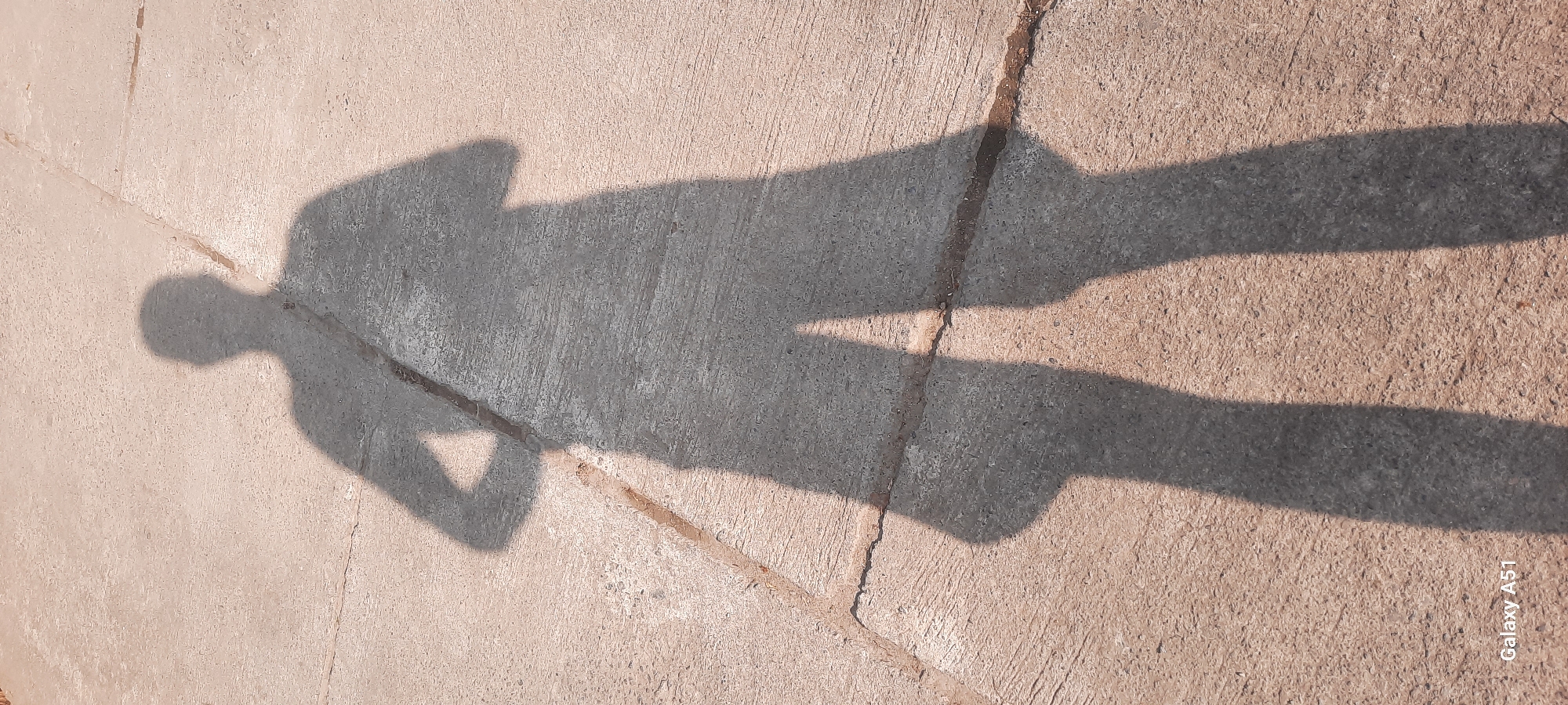 The image displays the shadow of a person standing on a concrete surface. The shadow is elongated and falls across the textured concrete, which is divided by faint lines. The lighting suggests it is daytime, with the sun casting a strong shadow. A portion of a bag strap is visible across the shadow's torso. The bottom left corner of the image includes the text "Galaxy A51." The location is identified as Jatitengah, Indonesia.