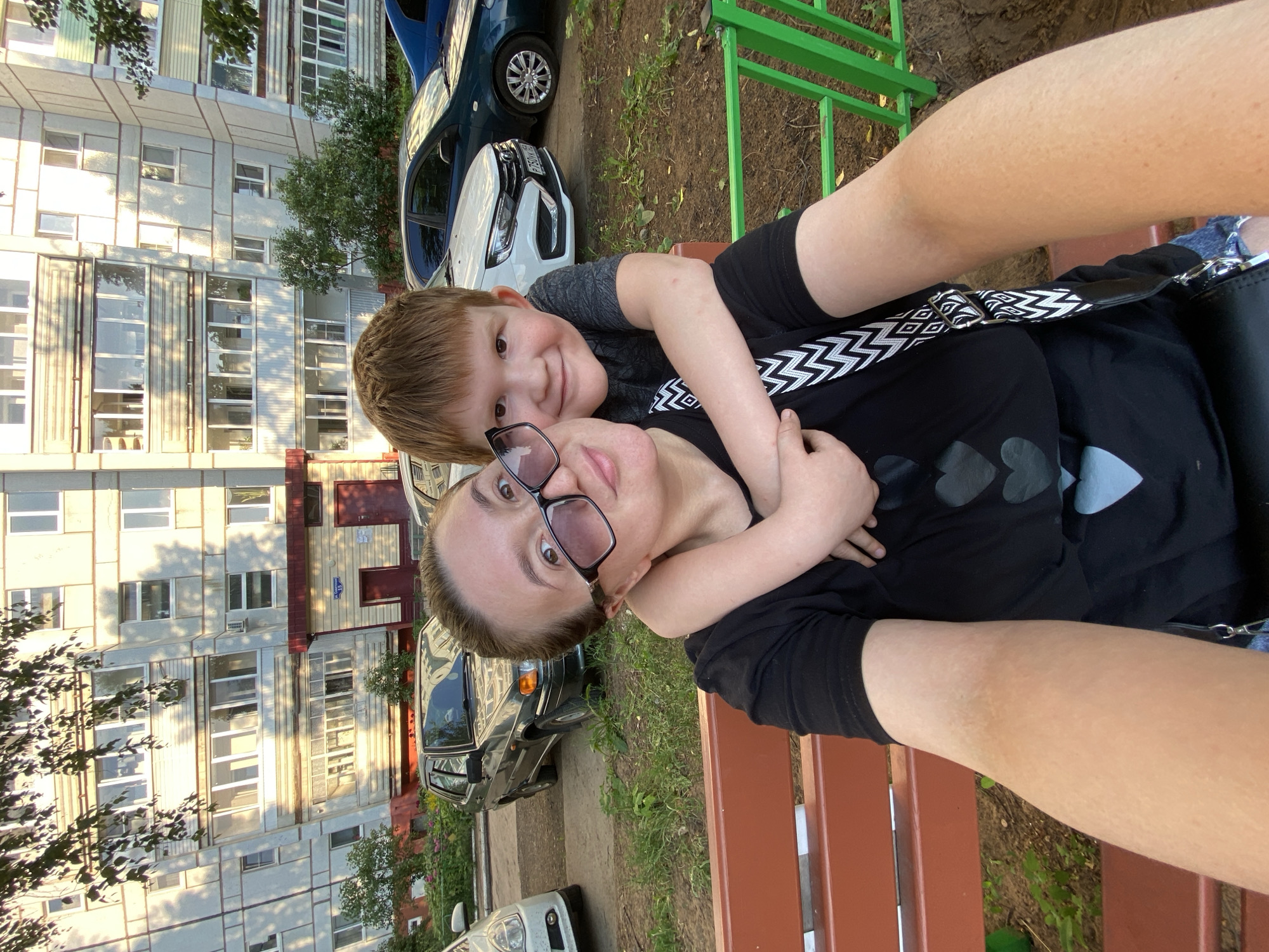 A young boy with reddish-brown hair hugs a woman from behind as they sit on a park bench. The woman, wearing large-rimmed glasses and a black t-shirt with a heart design, takes a selfie. Behind them, a grey apartment building with multiple balconies stands tall. Several cars are parked in front of the building, with a green fence and dirt ground in the foreground. The scene is lit by the warm glow of the sun.