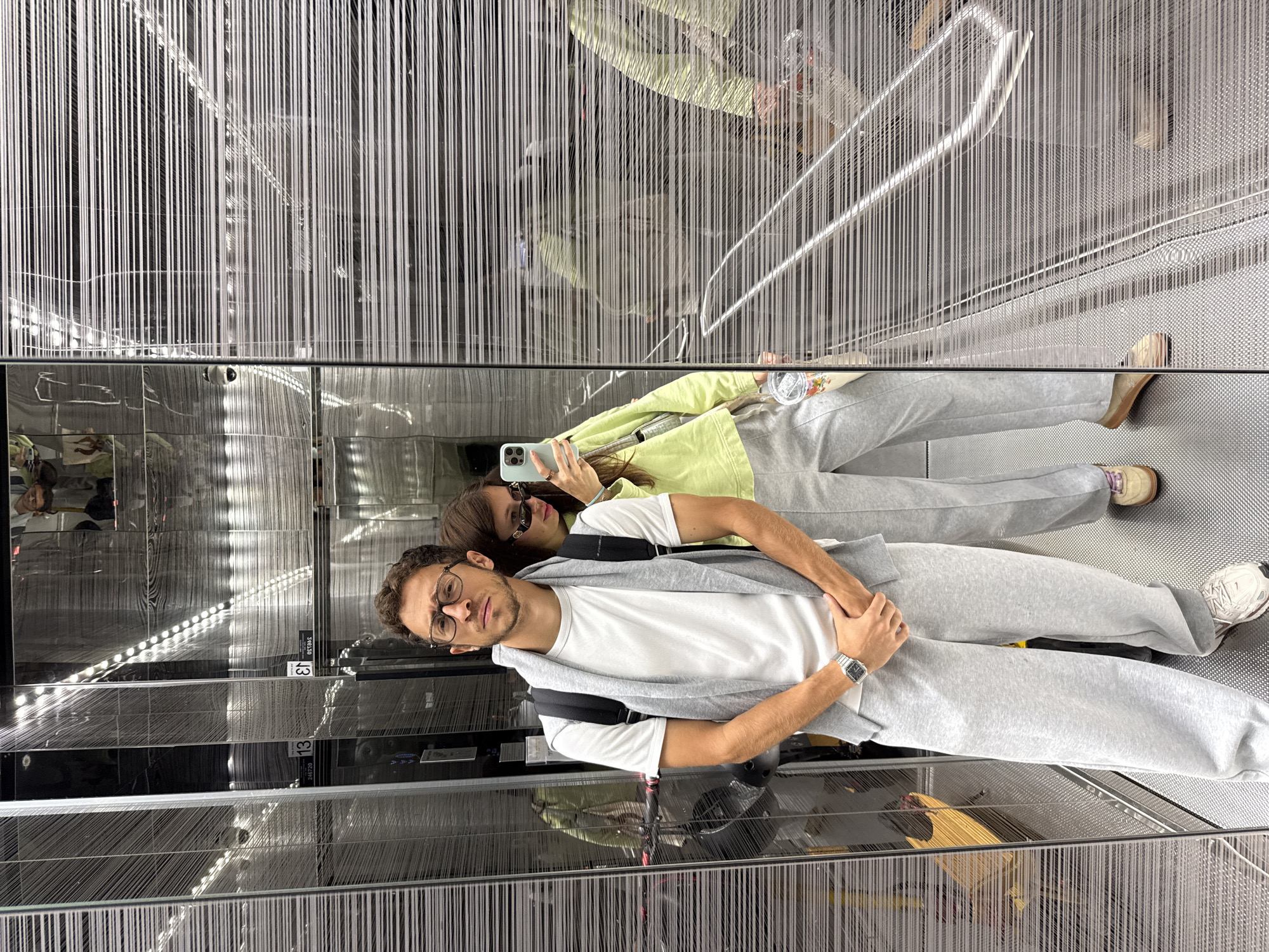 This media file captures a mirror selfie of two individuals, a man and a woman, inside what appears to be a modern building in Ramat Gan, Israel. The image is rotated 90 degrees counter-clockwise, showing the subjects lying horizontally across the frame.
In the foreground, on the left, a man with short brown hair and glasses is visible. He wears a white t-shirt, light grey sweatpants, and a grey hoodie or scarf draped over his shoulders, with a black backpack strap showing. A silver watch is on his left wrist. He looks directly at the camera with a neutral expression. Behind him, on the right, a woman is partially visible. She wears sunglasses, a light green jacket, and light grey sweatpants, holding a light blue smartphone to take the mirror selfie. Her face is mostly obscured. The man wears white sneakers with light grey accents, while the woman wears light brown/beige loafers.
The setting is highly reflective, suggesting polished metal or mirrored walls and ceiling within an escalator or moving walkway area. A striking feature is a dense pattern of thin, vertical white lines or strings that run throughout the scene, creating a sense of depth and a unique visual texture. The floor of the walkway has a textured, grid-like metallic surface. Reflections of the building's interior, including curved handrails and strip lighting (small white dots), are visible on the polished surfaces, creating a complex and somewhat disorienting visual effect.
The primary activity is the taking of a mirror selfie by the woman, capturing herself and her companion while in motion on the escalator or moving walkway. On the dark, reflective panels on the left side, fragmented text and numbers are visible in reflection, including "13", "P40720", and "P4384", which are likely facility indicators such as floor numbers or signage.