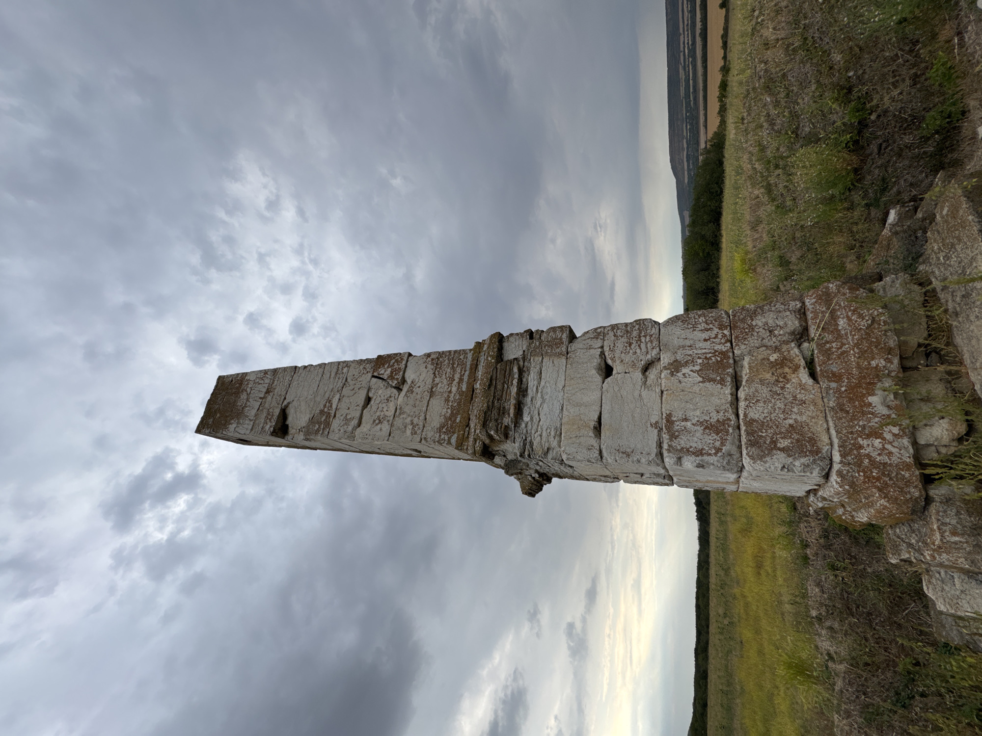 An ancient, weathered stone obelisk stands tall in a grassy field under a cloudy sky. The obelisk is constructed from large, rectangular stone blocks, some of which are cracked and show signs of erosion. A reddish-brown lichen or moss covers parts of the stone. The obelisk is positioned slightly off-center to the right in the frame, with its base resting on smaller, irregular stones and debris.
The field surrounding the obelisk is covered in tall, dry grass with patches of green and yellow. In the background, a line of trees forms a dark border against the horizon, beyond which are rolling hills and fields of harvested crops, visible in muted tones of yellow and brown. The sky is filled with a dense layer of gray clouds, with a sliver of lighter sky and subtle sunlight breaking through on the left side of the frame. The scene appears to be an open, rural landscape. This obelisk is believed to be located in the vicinity of Lesicheri, Bulgaria.
