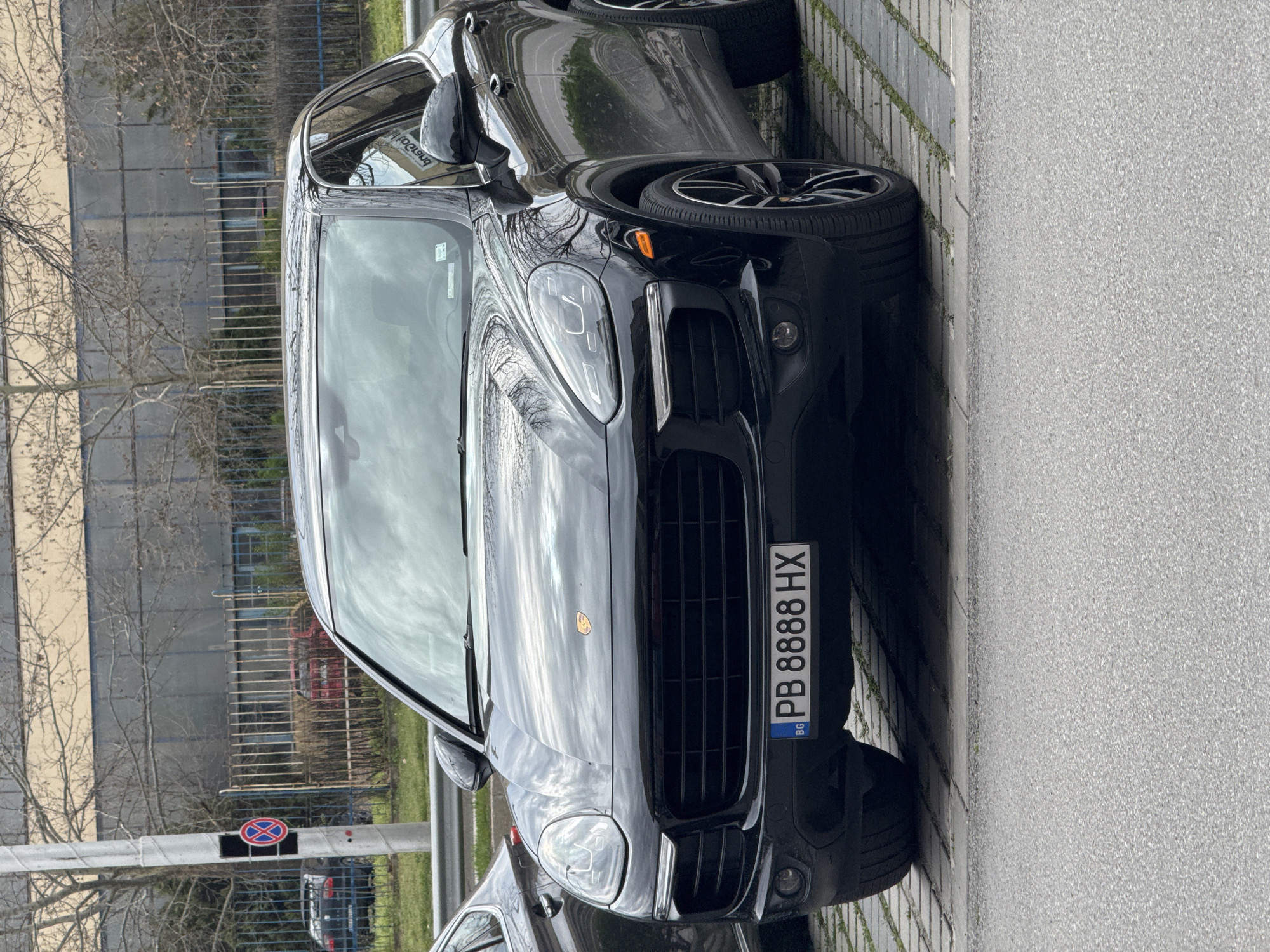 The image shows a black Porsche Cayenne parked on a paved surface, likely a parking area. The car's license plate reads "PB 8888 HX" and indicates registration in Bulgaria. A "No Stopping" sign is visible in the background, near a bare tree and a fence. Behind the fence appears to be a building, reflecting in the car's windshield.
Without further context or data, determining the precise location in Plovdiv is unfeasible.