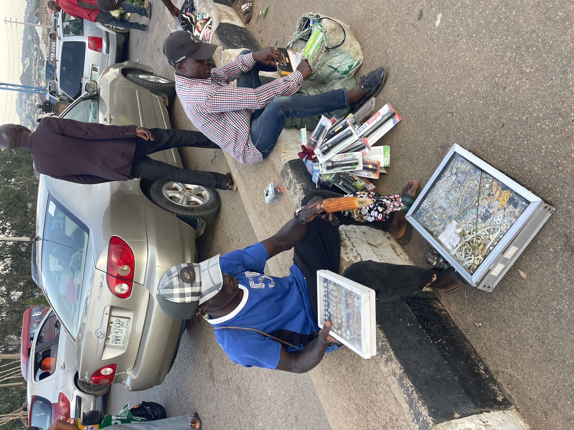 Two men are seen selling goods on the side of a street in Gura Top, Nigeria. The man on the left, wearing a blue t-shirt and a checkered cap, is seated on a concrete curb, holding a white tray filled with jewelry. To his right, another man, dressed in a red and white checkered shirt and blue jeans, is also seated and appears to be organizing items from a large green plastic bag, which contains various goods including flashlights. In front of him are more flashlights, some still in their packaging, and other assorted items. Behind the men, cars are parked along the street, indicating a busy urban environment. The setting appears to be a marketplace or roadside stall.