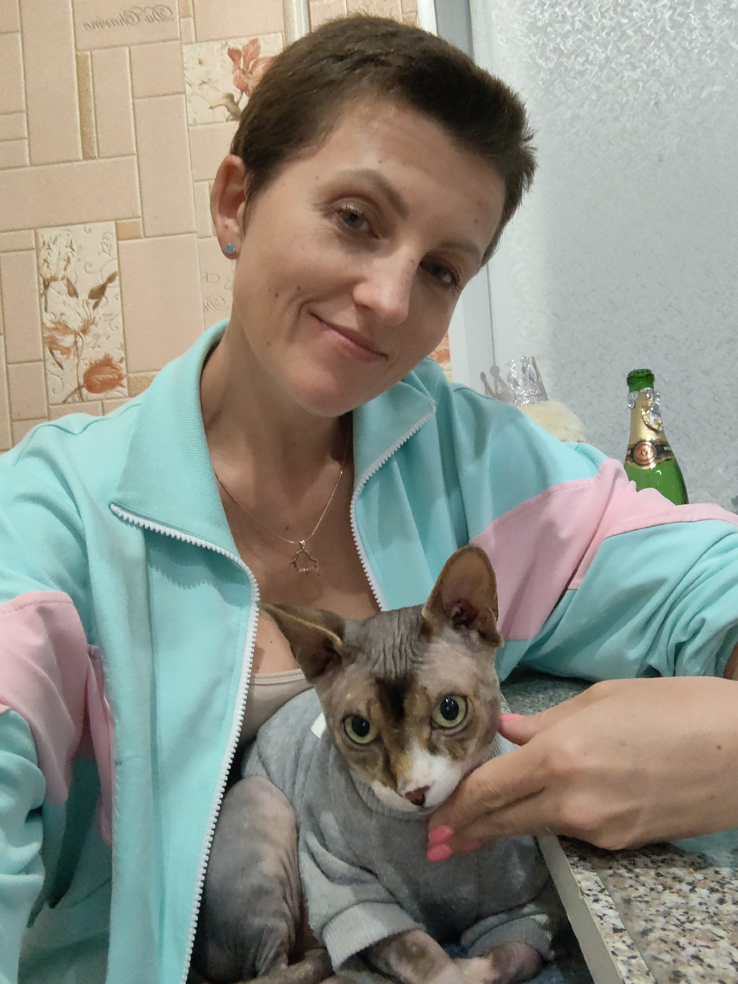 A selfie is taken by a woman with short brown hair and a light skin tone, wearing a light blue and pink jacket over a tan top. She has a delicate gold necklace with a triangular pendant. A light-skinned Sphynx cat, wearing a gray sweater, is held in her arms. The cat has large, green eyes and light brown markings on its face and body. The woman's right hand, with bright pink nail polish, is gently stroking the cat's cheek. In the background, a bottle of champagne with a gold foil wrapper and a green glass bottle is visible. The wall behind the woman is tiled with a pale pink floral pattern. The woman is smiling slightly, and the cat appears alert and attentive. The scene appears to be indoors, possibly in a home setting. There are no specific visual cues to indicate the location within the city of Myadzyel, Belarus.