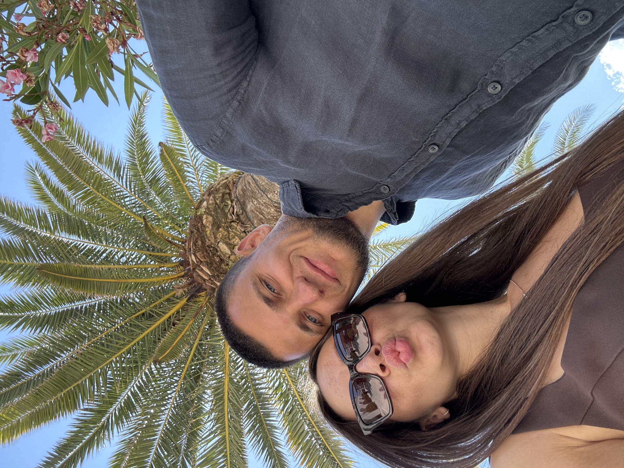 In Donja Lastva, Montenegro, a selfie captures a couple standing beneath a large palm tree with a bright blue sky as the backdrop. The woman in the foreground, wearing sunglasses and a brown top, puckers her lips for the camera. She has long, dark brown hair and is adorned with subtle earrings and a necklace. To her right, the man, sporting a dark button-up shirt, smiles warmly, his gaze directed at the camera. His hair is short and dark, and he has a light beard. The rough texture of the palm tree trunk is visible behind them, and its green fronds spread out overhead, with a few pink flowers from a nearby plant visible at the top right of the frame.