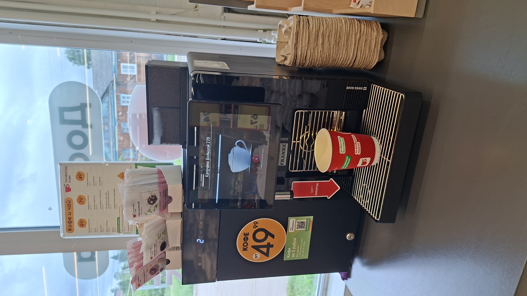 The image shows a self-service coffee machine, a Franke A300, located inside what appears to be a convenience store or a small cafe, possibly at the Pyaterochka supermarket in Novoilinsky, Russia. The coffee machine is black and has a digital touchscreen display showing "Капучино Большой 270" (Cappuccino Large 270). There is a red paper cup with a lid placed on the drip tray, waiting to be filled. Above the machine, a sign with "Кофе и чай" (Coffee and tea) and prices like 99, 79, and 49 is visible, along with product packaging for tea bags. To the left of the coffee machine, there is a circular orange sticker with "КОФЕ ОТ 49⁹⁹" (COFFEE FROM 49⁹⁹) and a QR code. The environment is indoors with a window in the background showing trees and buildings, suggesting it is daytime. The overall scene is clean and organized, focusing on the coffee machine and its offerings.