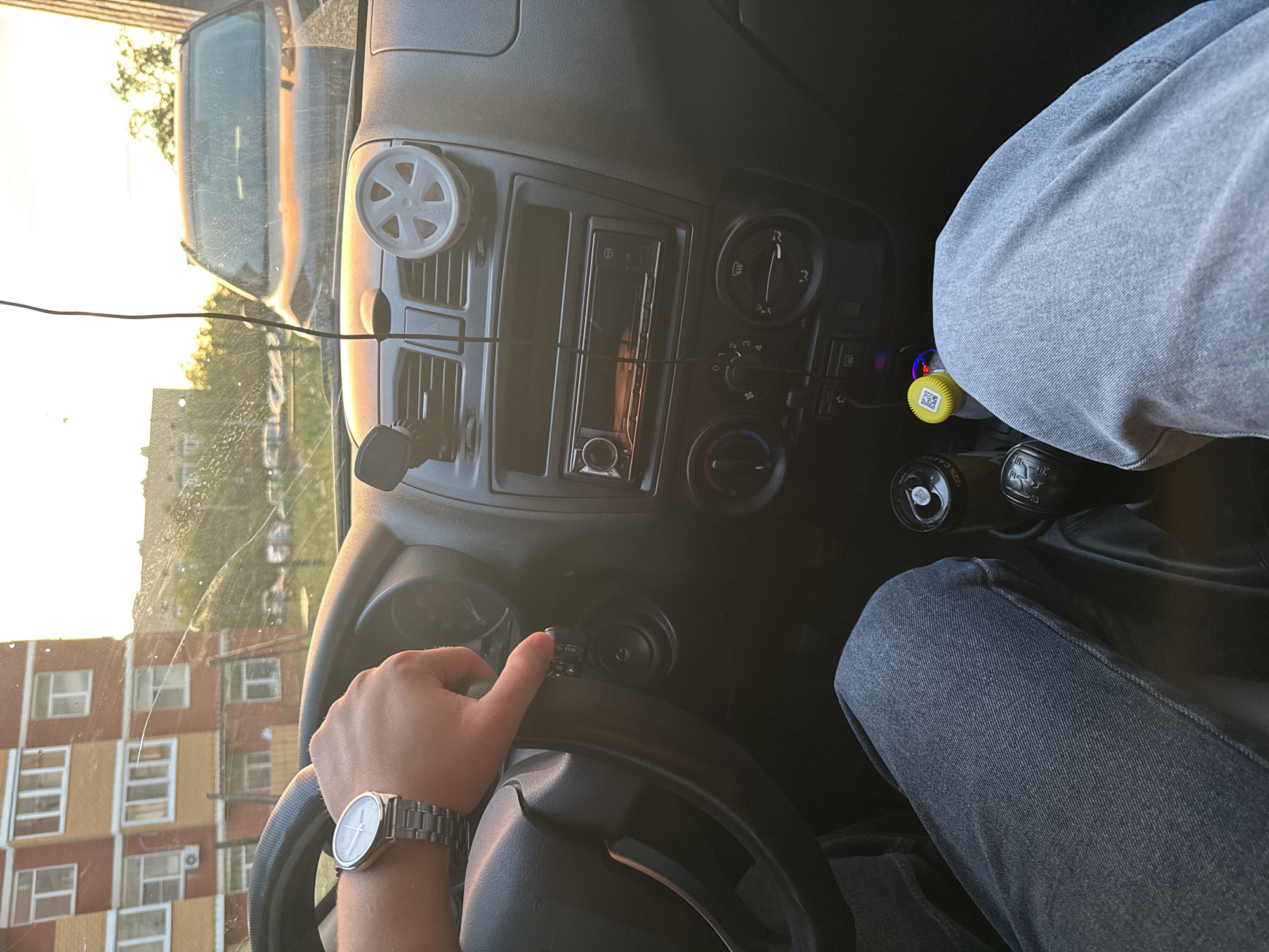 A view from inside a car in Perm, Russia, shows a person driving. The driver wears a silver wristwatch on their left hand. The car's dashboard is black and contains a radio, climate control dials, and air vents. A cracked windshield is visible. Outside the car, a light-colored building with multiple windows is seen. The gear stick is on the right of the driver with an unusual yellow-topped object plugged into the cigarette lighter. The car is a Lada.