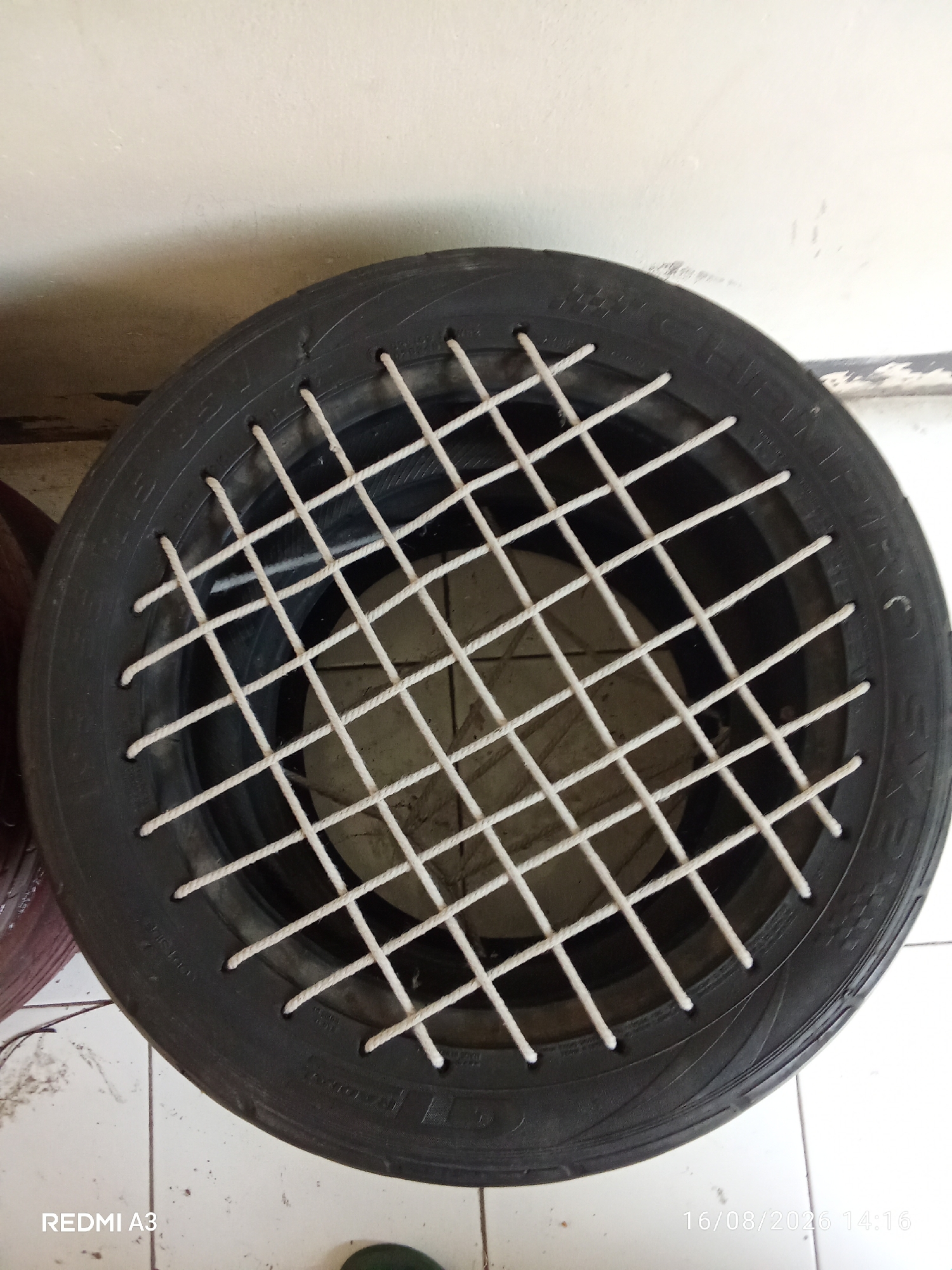 A tire has been repurposed as a planter. The tire is black and sits on a tiled floor. A white rope grid has been laced through holes in the tire wall, creating a criss-cross pattern over the opening of the tire. Inside the tire, a light-colored, circular object is visible. The background consists of a light-colored wall and a tiled floor. A watermark in the bottom left corner reads "REDMI A3" and in the bottom right corner reads "16/08/2026 14:16". The image was taken in Surakarta, Indonesia.