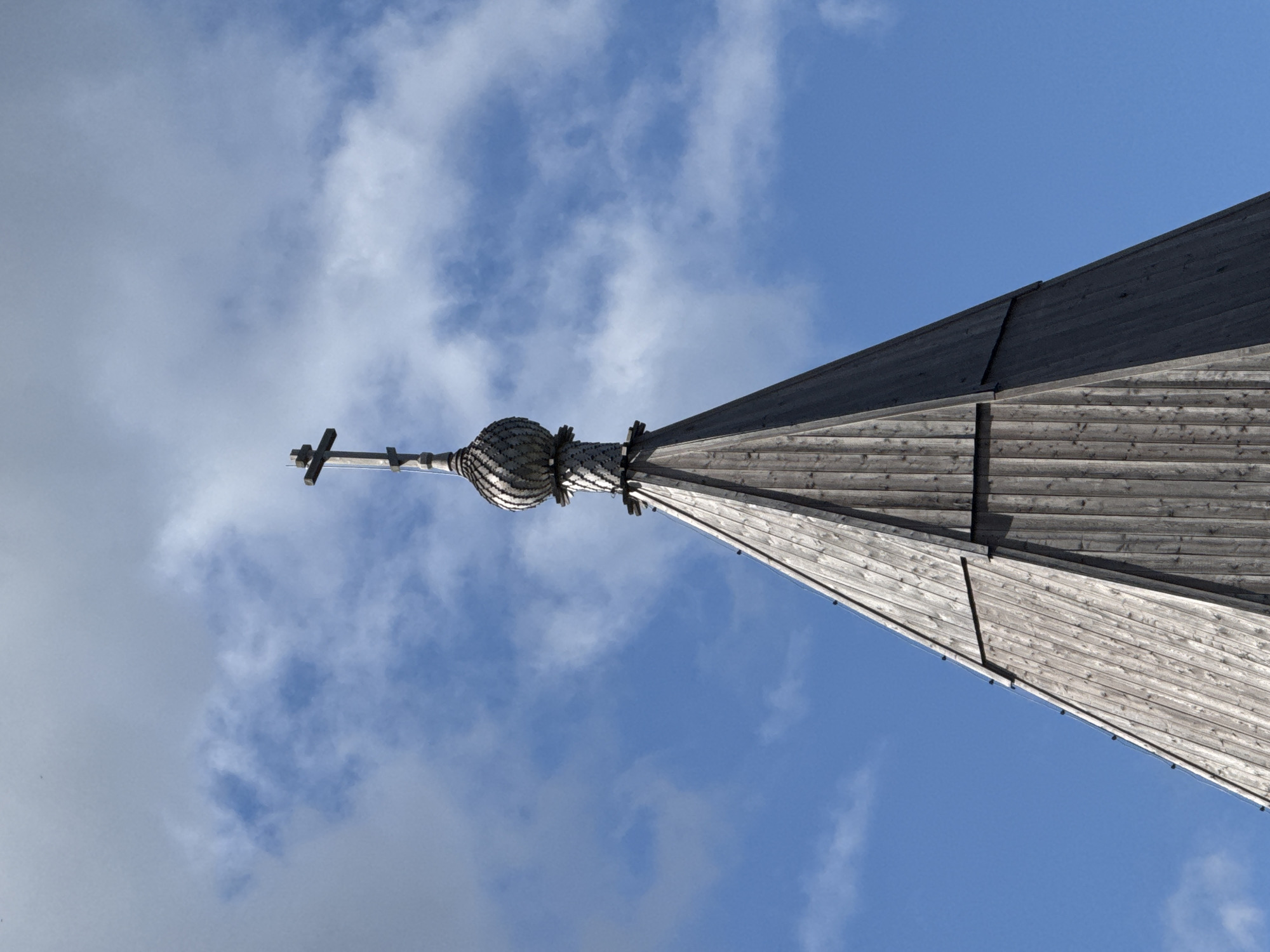 This is a low-angle, eye-level, medium shot of a wooden church spire topped with a cross against a bright blue sky with scattered white clouds. The spire is constructed from vertical wooden planks, weathered to a gray tone. A bulbous, layered dome sits below the cross, also made of wood. The cross itself is a traditional Orthodox Christian cross, with a horizontal bar and two smaller diagonal bars. The image appears to be taken outdoors in a rural setting, likely the village of Gora in Khokhlovskoe rural settlement, Russia, given the architectural style of the church spire. There are no people or animals visible in the frame. The lighting suggests it is daytime, with the sun illuminating the spire from the side, creating some shadows on the wooden planks. The overall impression is one of serene, religious architecture set against a natural backdrop.