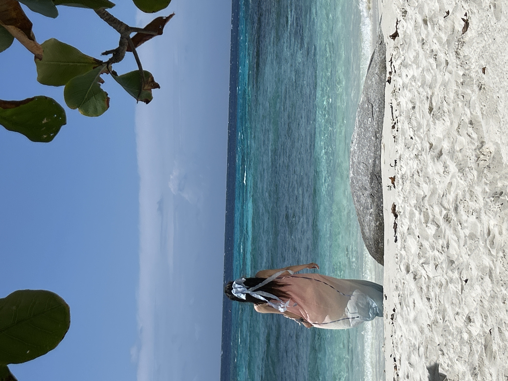 A person wearing a light-colored dress with blue ribbon accents in their hair stands on a sandy beach, facing a clear turquoise ocean under a blue sky. The water exhibits multiple shades of blue and green, with gentle waves reaching the shore. A large, gray rock is situated on the beach to the right of the person, and scattered driftwood and leaves are visible on the sand. Tree branches with green and brown leaves frame the top of the image. The scene is consistent with a location in Rawai, Thailand, known for its beautiful beaches and clear waters.