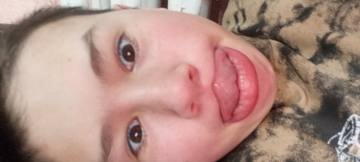 The image presents a close-up view of an individual's face, rotated approximately 90 degrees clockwise, in an indoor setting within Красный Маяк, Russia. The subject's face occupies the majority of the frame, featuring fair skin with visible capillaries on the cheeks. Dark brown hair is visible at the top and left edges of the head. Both brown eyes are open and directed towards the viewer. The nose is central, and the mouth is open with the tongue extended outward, appearing pink and moist. The individual is wearing a garment with a mottled light brown and black pattern, visible on the right side of the frame. A small, indistinct light-colored object with dark markings is partially visible in the bottom right corner. The background is softly blurred, showing light-colored fabric in the upper left and a vertical, light-toned surface towards the top-center.