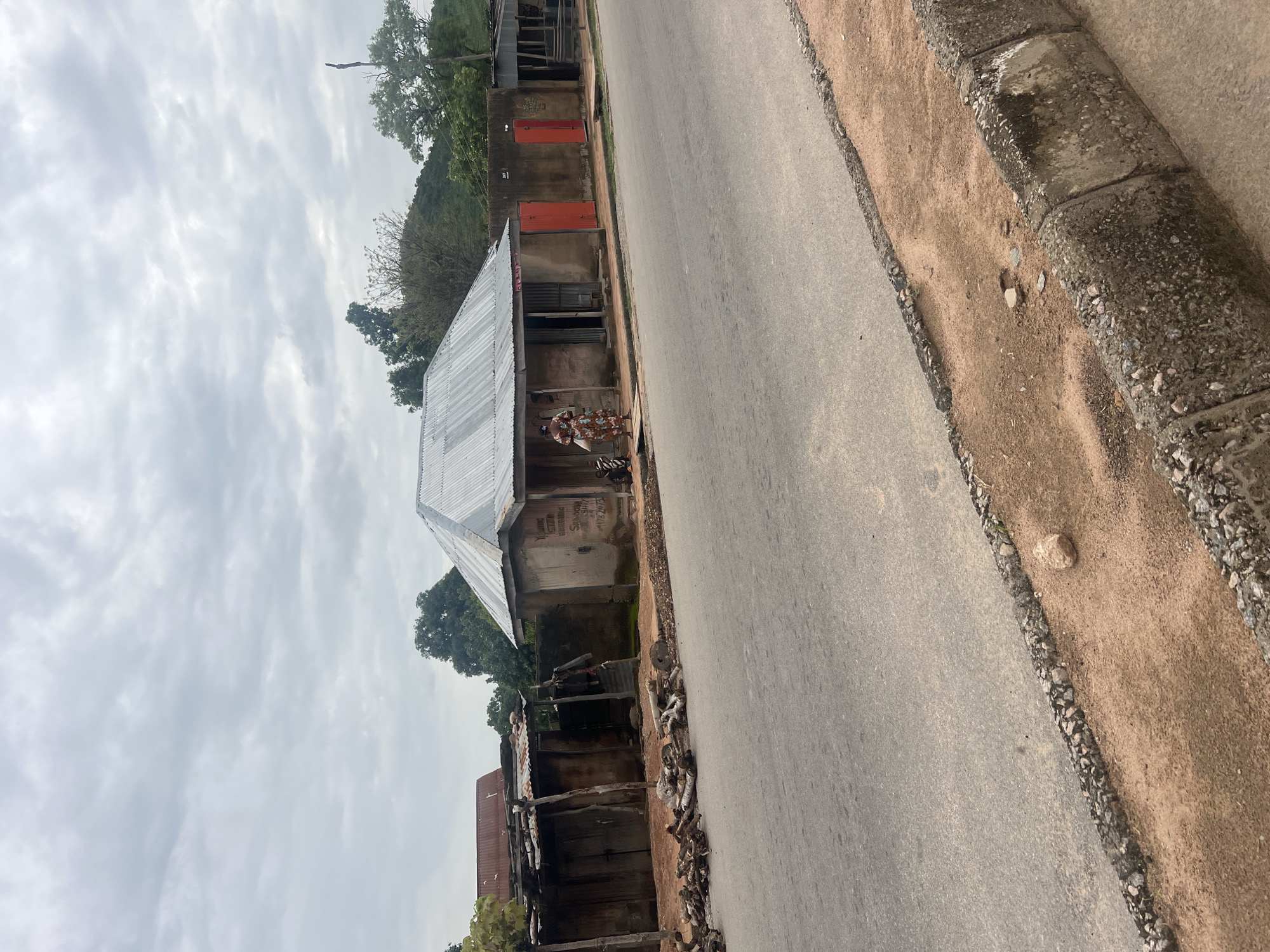 The image depicts a street scene in Kona Garu, Nigeria. A person is standing in front of a building with a corrugated metal roof. The building appears to be a shop or dwelling. Next to it, another structure with a similar roof is visible, and further down the street, more buildings and trees can be seen. The road is paved and runs alongside a sandy embankment with a stone border. The sky is overcast, suggesting cloudy weather. The overall scene appears to be a typical day in a Nigerian town or village. There is some text visible on the wall of the building in the foreground, but it is illegible.