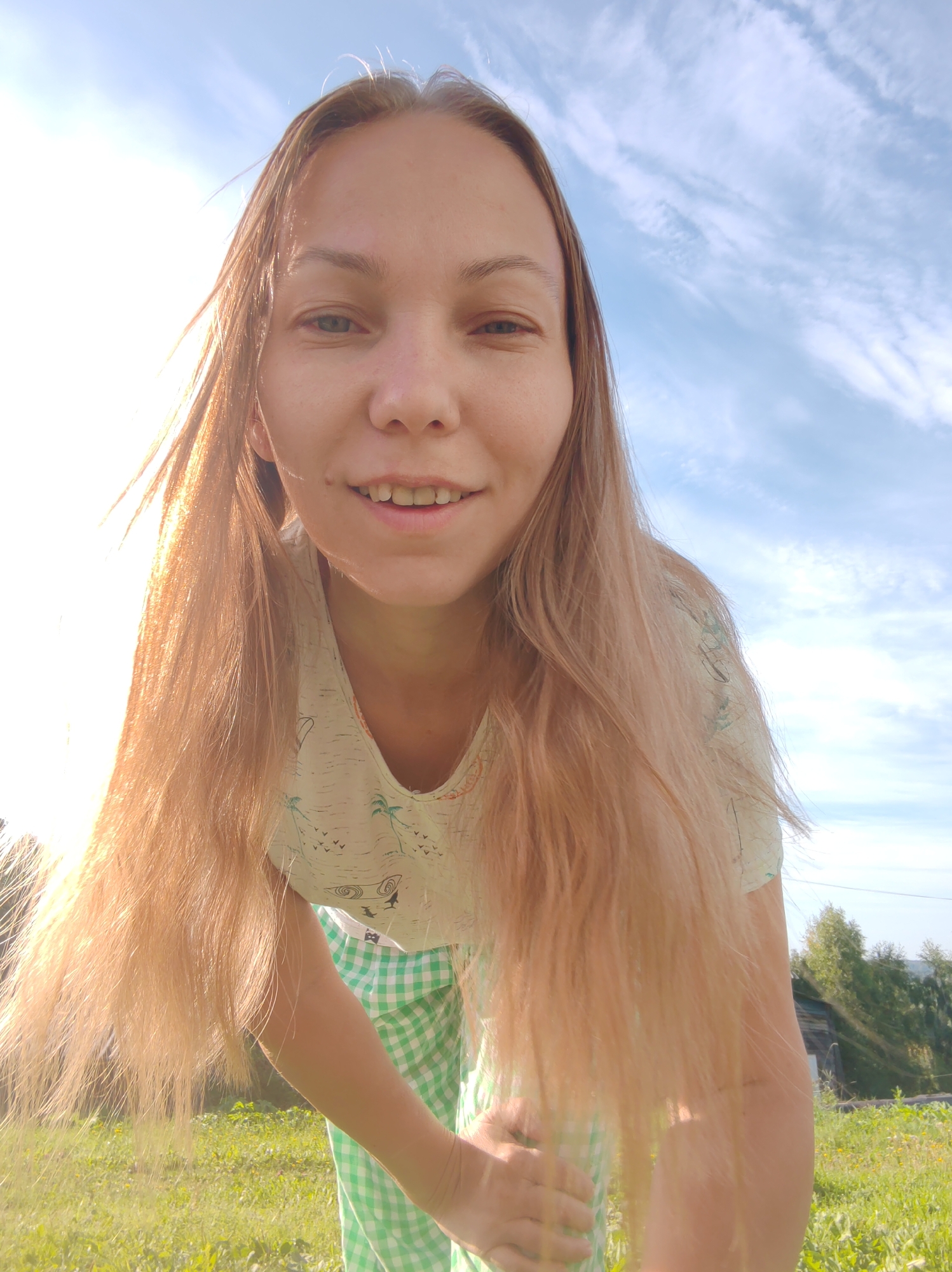 A young woman with blonde hair is posing for a selfie outdoors. She is leaning forward, looking at the camera with a slight smile. She is wearing a light-colored t-shirt with a tropical print and a green and white checkered skirt. The background shows a grassy field with some trees and a cloudy blue sky. The scene appears to be in a rural area, potentially in the city of Chuchlemlia, Russia, given the wooden building visible in the distance. The lighting suggests it is daytime, possibly late afternoon due to the warm glow.