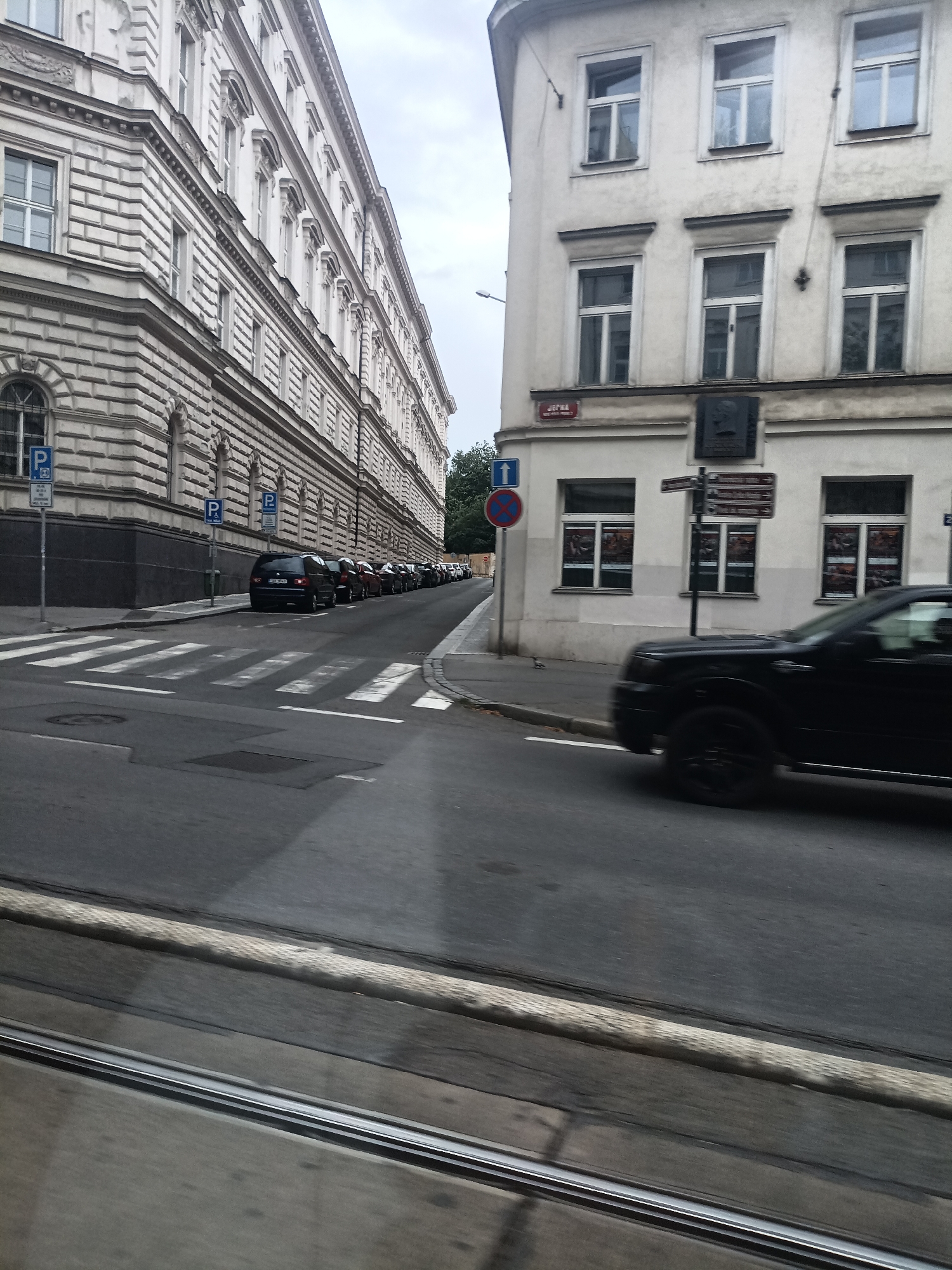 The image depicts a street scene in Prague, Czechia. On the left, a grand, ornate building with multiple windows and decorative architectural features lines the street. A row of parked cars is visible along the sidewalk, some with parking signs. On the right, another building with a lighter facade and several windows stands. A black SUV is in motion, blurring as it drives across the street. Traffic signs are visible, including a blue sign with a white "P" indicating parking and a circular sign with a red border and a diagonal red bar, likely a prohibition sign. A pedestrian crossing is marked on the road. The sky appears overcast, suggesting a cloudy day. The overall impression is a typical urban street, possibly in the morning or afternoon, given the lighting.