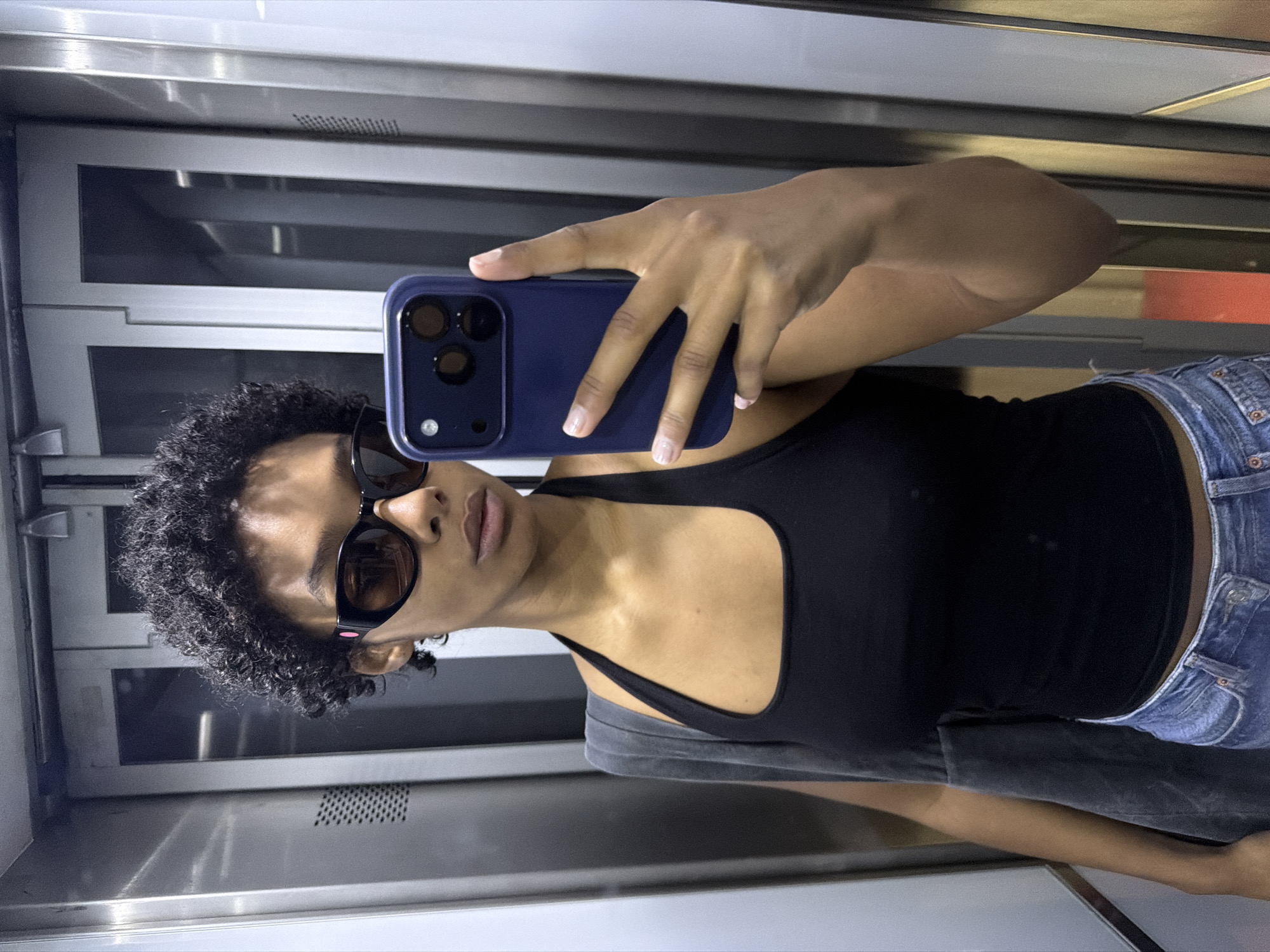 The image shows a young woman with dark, curly hair and dark sunglasses taking a selfie in an elevator. She is wearing a black tank top and blue jeans. A dark grey bag is slung over her left shoulder. The elevator has brushed metal walls and doors with vertical ribbed panels. The lighting is somewhat dim and reflective. There is no indication that the image was taken in Paris, France.
