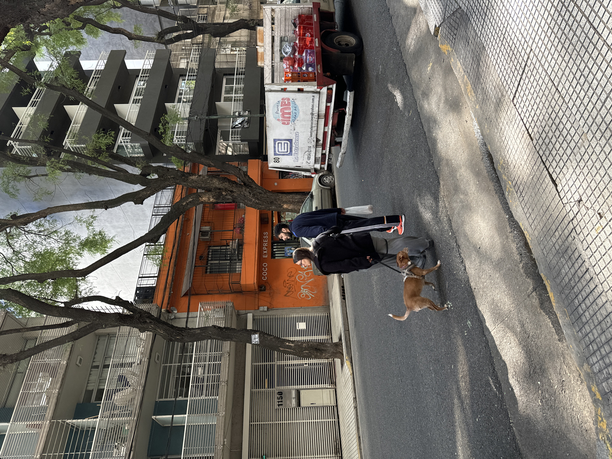 The image depicts a street scene in Buenos Aires, Argentina. A person wearing a black jacket and glasses is walking a brown dog with a leash. Another person in a blue jacket and orange shoes walks nearby. They are on the asphalt road next to a curb with a grid pattern. A truck with the brand name "Cimes" on the side is parked on the road. The truck is loaded with crates of what appears to be beverages. Buildings line the street, one of which is painted orange with the words "COCO EXPRESS" and graffiti on the facade. The address "1150" is visible on another building. Trees with bare branches are present along the street.