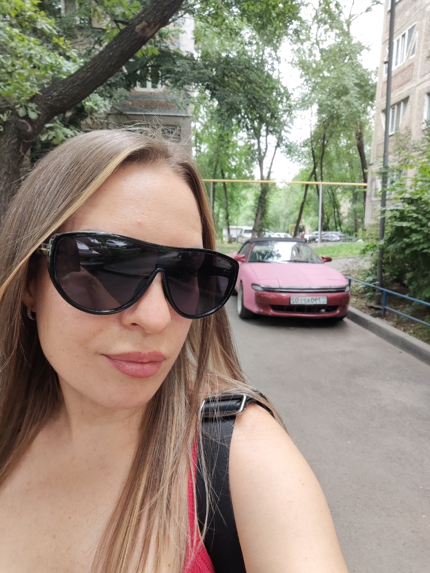 A medium shot shows a fair-skinned woman with blonde hair and sunglasses in the foreground, taking a selfie. She is looking downwards and slightly to her left. She wears a pink top and a black bag strap is visible. In the background, a pink car is parked on a street. Trees and a building are visible in the background. The location is a residential area in Almaty, Kazakhstan.