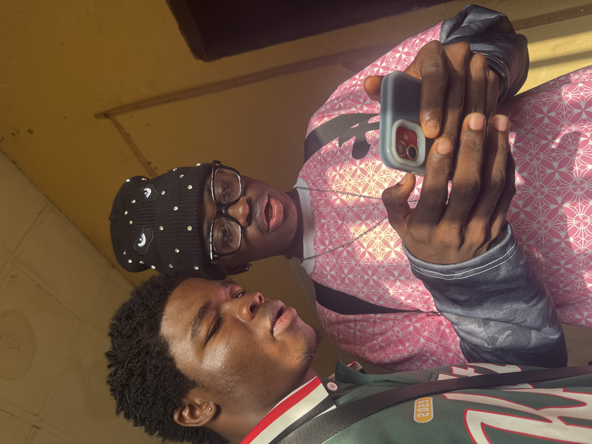 Two African men are visible in the frame. One is lying down, eyes closed, wearing a green shirt with white and red stripes and the number "2025" on the sleeve. The second man is standing above him, holding a cell phone in both hands, taking a picture or recording a video. He is wearing a black hat with silver embellishments, glasses, and a pink patterned shirt. The photo is taken indoors, with yellow walls in the background. The image suggests the men are in Ihiagwa, Nigeria based on the prompt.
