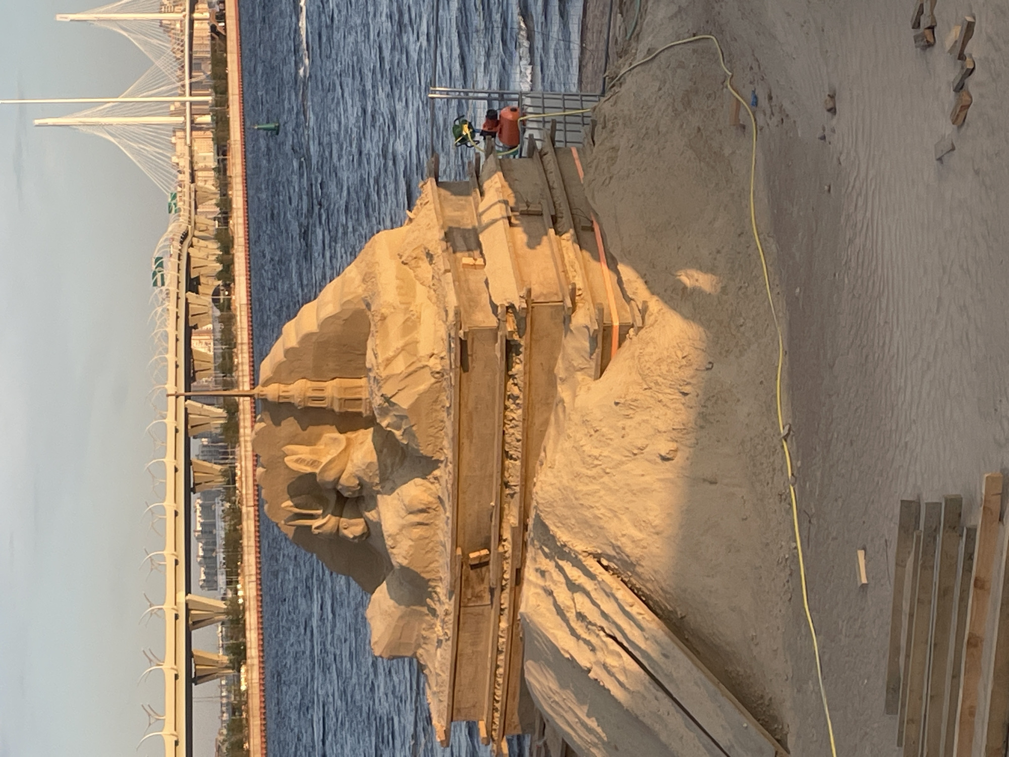 A sand sculpture, resembling a tiered structure with a small tower and animal figures on top, is situated on a sandy embankment next to a body of water. The sculpture is illuminated by the warm, low light of the sun, casting long shadows. In the background, a large modern bridge spans across the water, with city buildings visible in the distance. A green buoy floats in the water near the shore. Equipment, including a red water pump and a metal rack, is present near the right side of the sculpture. Wooden planks and loose bricks are scattered on the sand in the foreground. The sky is clear with a hint of pale blue at the horizon.