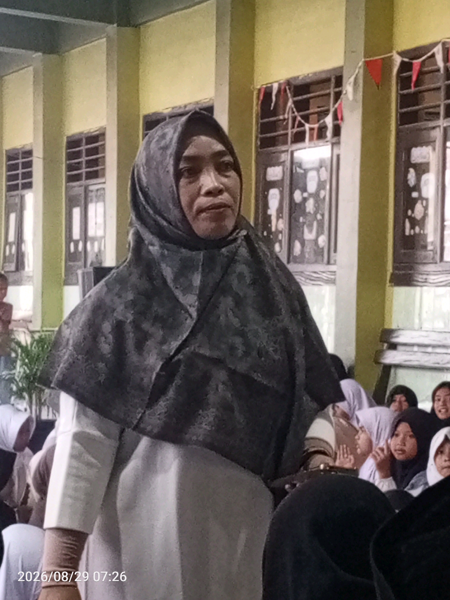 The image captures a medium shot of a woman wearing a dark patterned hijab and a light-colored outfit. She is looking to her right with a neutral expression. In the background, several other individuals are visible, many wearing hijabs, suggesting a gathering or event. The environment appears to be outdoors, with a building featuring yellow walls and windows with barred panes. Red and white triangular flags are strung across the top of the windows. A plant is visible on the left side of the frame. A timestamp "2026/08/29 07:26" is overlaid in the bottom left corner of the image. The scene is likely situated in Tanjungan, Indonesia, given the provided context.