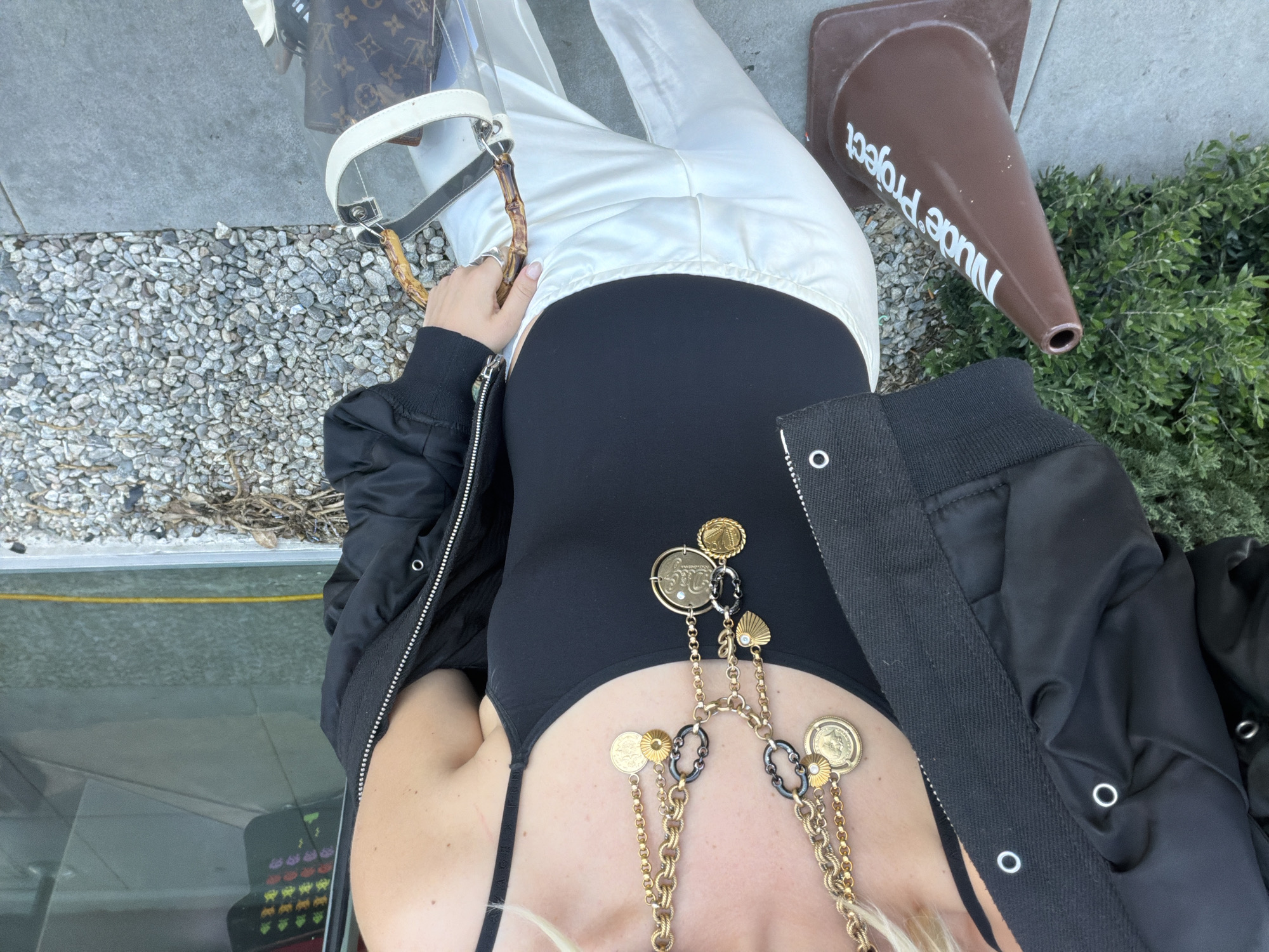 The image depicts a person wearing a black bodysuit, white pants, and a black bomber jacket, with a long gold chain necklace. They are holding a Louis Vuitton handbag with a clear exterior and bamboo handle. A brown traffic cone with the words "Nude Project" printed on it is visible in the background, along with some greenery. The ground is covered in small pebbles. The setting is outdoors in West Hollywood, United States of America.