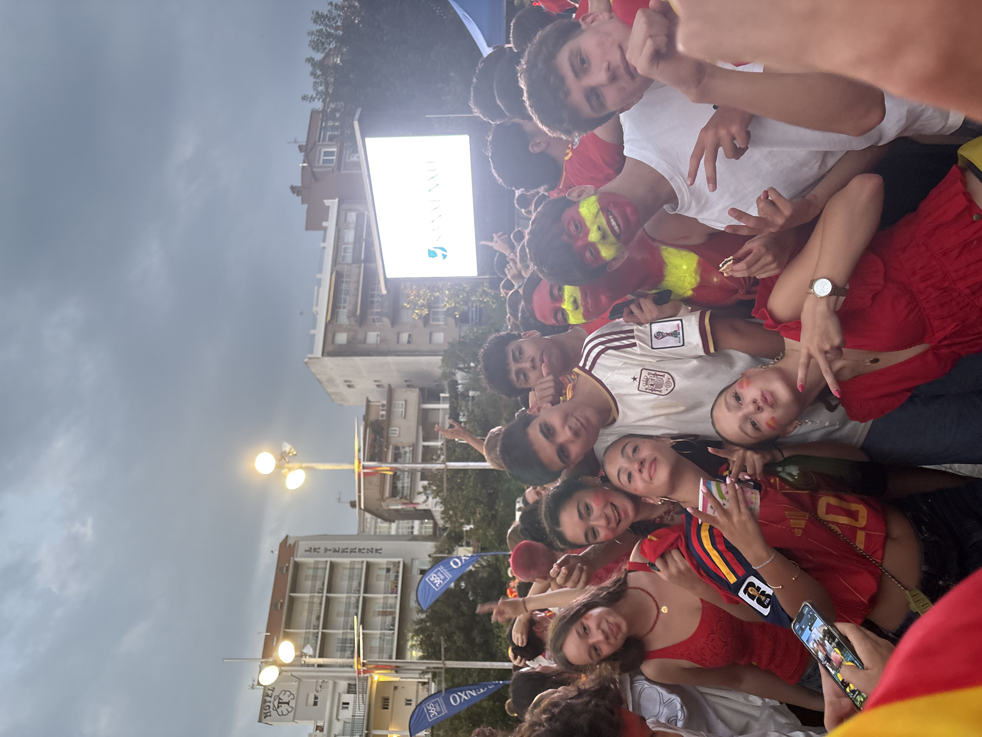 In Sanxenxo, Spain, a group of young people, some with their faces painted in the colors of the Spanish flag, are gathered outdoors, appearing to watch an event on a large screen. They are dressed in casual clothing, many in red and yellow, suggesting a celebratory or patriotic atmosphere, likely related to a sporting event. The background features buildings, streetlights, and flags, indicative of a public square or outdoor venue. The sky is overcast, suggesting it could be late afternoon or early evening. The individuals are smiling, posing for the camera, and some are holding drinks or food.