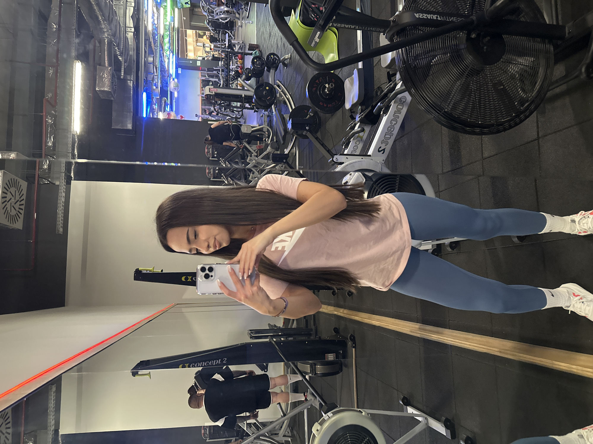 A full-length shot captures a woman in a gym setting, taking a selfie using a smartphone. She is dressed in a pastel pink t-shirt and blue leggings, coupled with white sneakers. Her long, dark hair cascades down her back.
The gym backdrop includes various exercise machines, such as a stationary bike and rowing machine, and other gym-goers in the distance. A mirror reflects the gym environment, creating a mirror image of the scene and the woman taking the selfie. The flooring is dark and made of rubber, indicating it is a gym or fitness facility. The location and city are not discernible from the image.