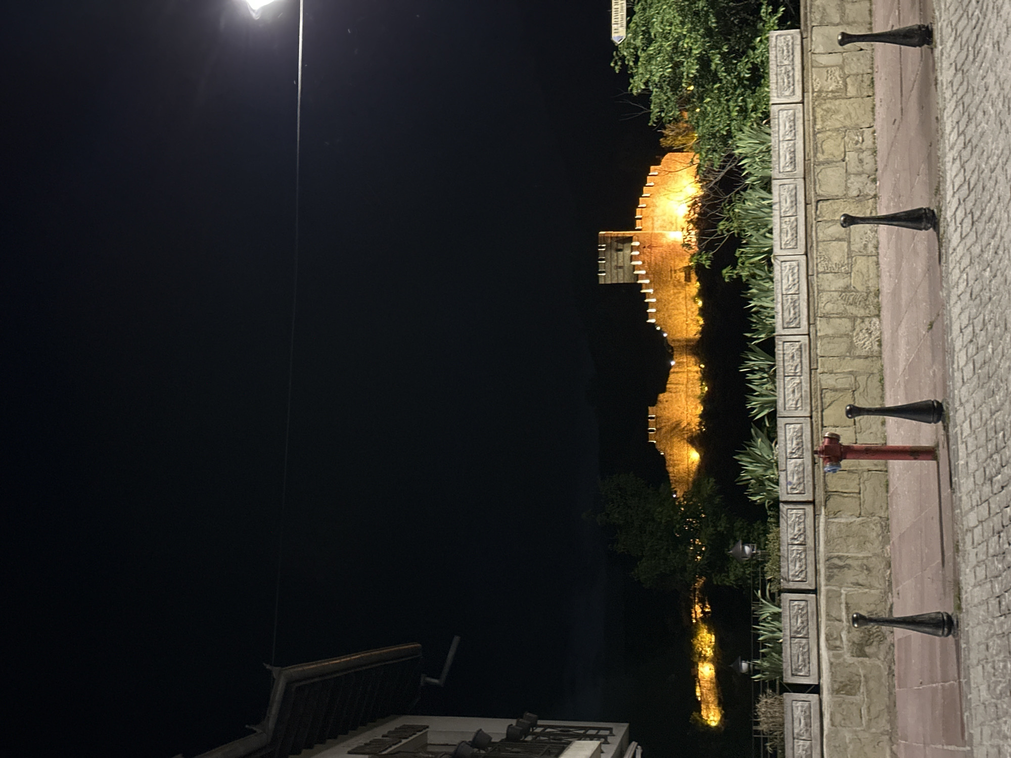 At night in Veliko Tarnovo, Bulgaria, a fortress wall is illuminated, its reflection visible in the water below. A street lamp stands to the side, and a building is seen in the foreground. A stone wall with decorative planters runs along the edge of the walkway, punctuated by bollards and a red fire hydrant. The walkway is paved with cobblestones.