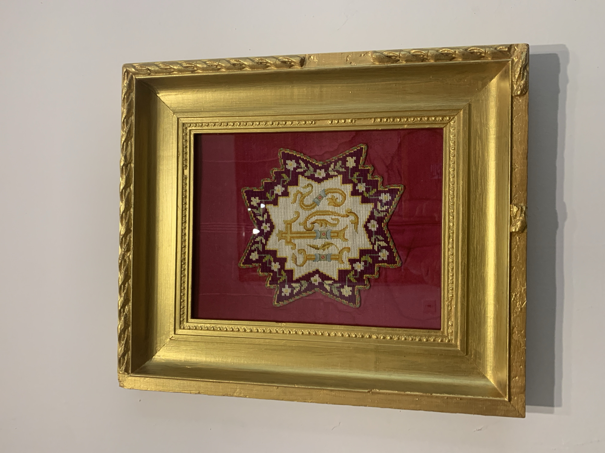 A framed needlepoint artwork is displayed on a white wall. The artwork is star-shaped and features a monogram with the letters "JHS" in gold thread, surrounded by a border of purple and white stitching. The needlepoint is mounted on a red fabric background and encased in a glass frame. The frame itself is ornate, with a gold finish and intricate detailing along the edges, including a rope-like pattern and smaller decorative elements. The gold finish shows some signs of wear and tear, with chips and scuffs visible, particularly along the bottom edge. The artwork is hung at eye level, with a shadow cast on the wall beneath it, suggesting it is lit from above. The overall presentation indicates a preserved piece of decorative art. There is no visible information to place this image in Kutaisi, Georgia.