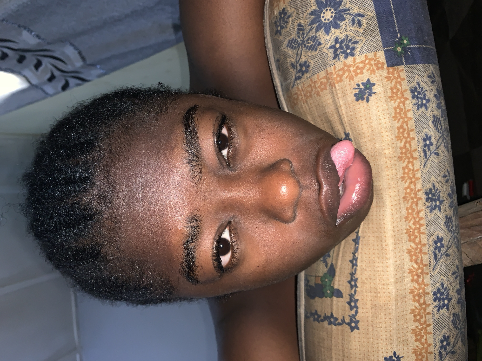 A close-up, high-angle shot depicts a dark-skinned individual lying on a patterned cushion or mattress. The person's head is slightly tilted. Their mouth is partially open with their tongue protruding slightly. The background includes a patterned covering. The location is Rafin Par, Nigeria.