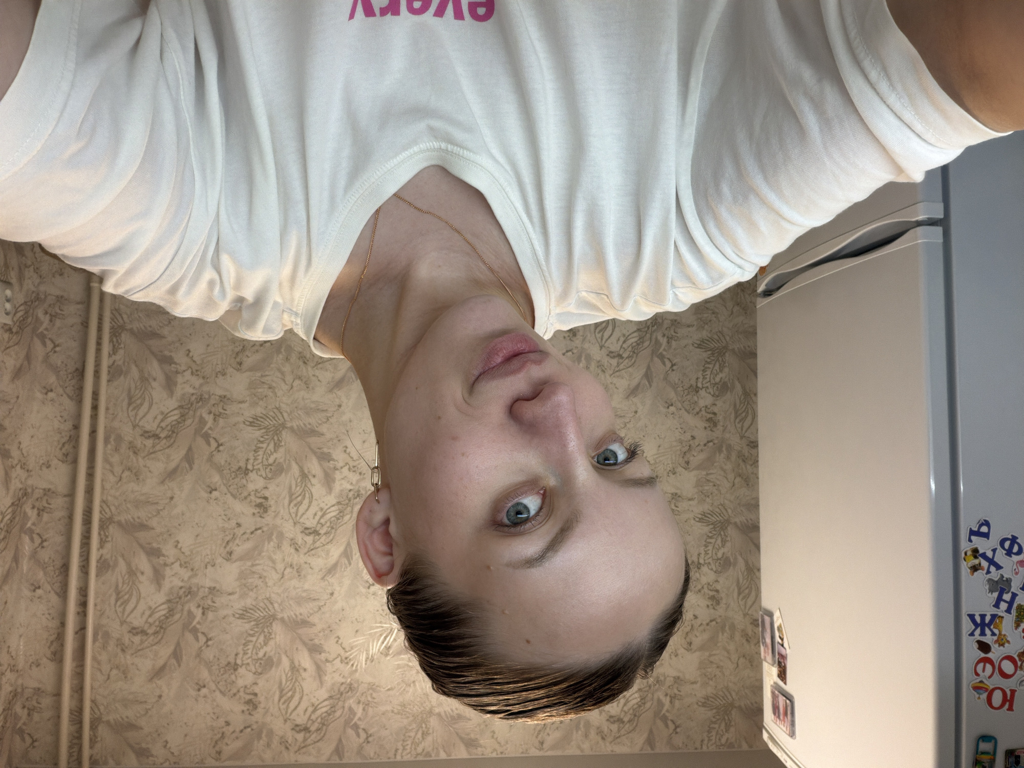 The image shows a woman with light skin, short brown hair, and blue eyes, wearing a white t-shirt and a gold necklace. She is looking up at the camera. Her left ear is visible, and she is wearing an earring. The background consists of a patterned wallpaper and a white refrigerator with magnets on it. The location is potentially Reshetnikovo, Russia, but this cannot be verified from the image.