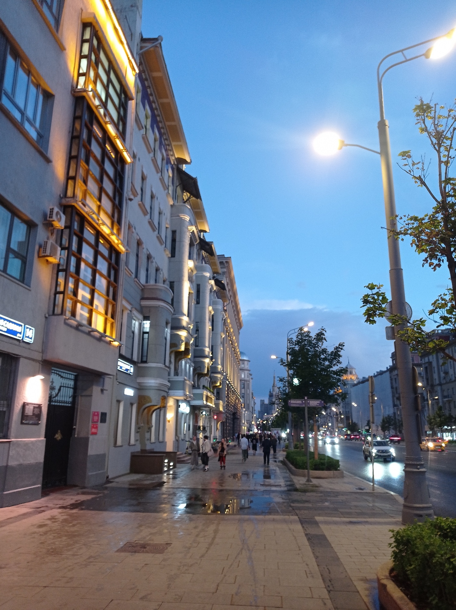 The image depicts a street scene in Moscow, Russia, during twilight. The perspective is from a sidewalk, looking down a wide street lined with ornate, multi-story buildings. The architecture of the buildings features decorative balconies and illuminated windows, suggesting a classical or art nouveau style. The sky is a deep blue, transitioning from the day.
On the left side of the street, a building with large, grid-patterned windows is prominent, with warm yellow lights illuminating its facade. A sign with Cyrillic lettering and the number "14" is visible on this building. To the right of this building, the sidewalk is wet, with reflections of lights from the buildings and streetlamps. Several people are walking on the sidewalk, some in groups, their figures somewhat blurred by the low light and motion.
The street itself has multiple lanes of traffic. Several cars are visible, their headlights and taillights creating streaks of light due to the long exposure or low light conditions. The street is also lined with trees and streetlamps, which are casting pools of light on the wet pavement. In the distance, more buildings and lights are visible, including what appears to be a dome or spire on one of the distant structures.
The overall impression is that of a well-lit urban street at dusk, with wet conditions on the ground suggesting recent rain. The presence of pedestrians and cars indicates a typical evening in a bustling city.