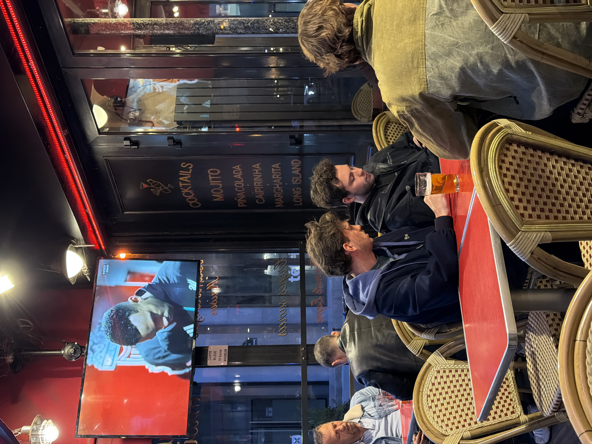 Here is a detailed description of the image:
**Setting:** The image captures a scene inside a Parisian café or bar. The presence of classic wicker chairs, red tables, and cocktail menu signage indicates this type of establishment. The reflection in the glass windows and doors suggests an urban environment, possibly in Paris, France. A TV displaying a sports game can be seen in the background.
**People:** The photograph features several people. Most are seated at tables, potentially engaged in conversation or watching the television.
**Objects:** Notable objects in the image include:
* Wicker chairs, a common design found in Parisian cafés.
* Red-topped tables.
* A glass of beer in a man's hand.
* A television displaying a sports game, likely soccer (football).
* A sign with a list of cocktails, including "Mojito," "Pina Colada," "Caipirinha," "Margharita," and "Long Island."
* A menu indicating that "Carte Bleue" (a debit card) is accepted with a minimum of 10 Euros.
**Actions and Interactions:** The people in the photograph are primarily seated and seem to be engaging in leisurely activities, such as drinking and watching television. The interaction between people at the tables is not directly captured.
**Environment:** The environment is indoors, with artificial lighting. The reflections in the glass indicate that it is likely evening or night. The overall impression is that of a relaxed social atmosphere in a typical Parisian bar.