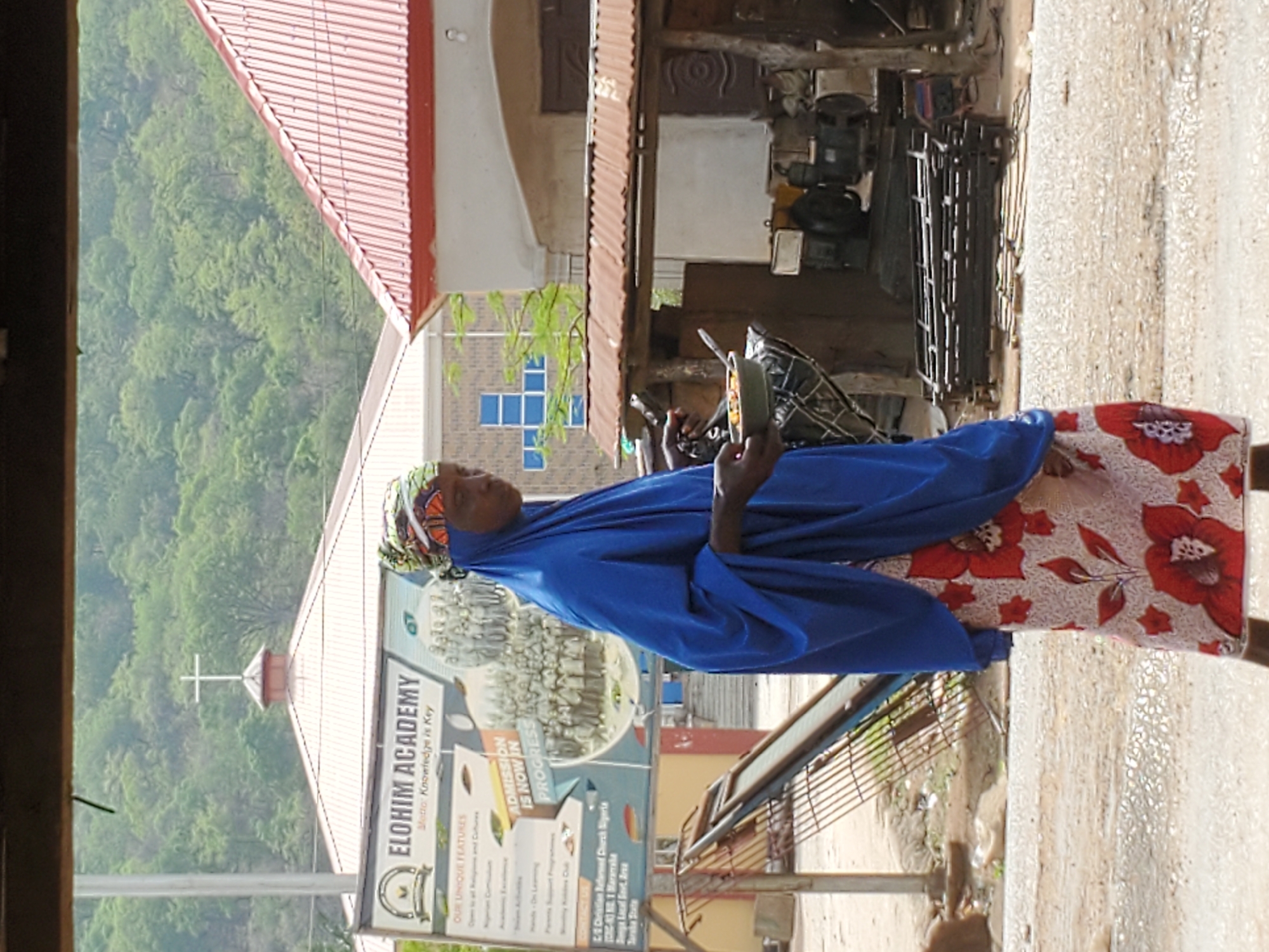 This image, captured in Mararaba, Nigeria, depicts a daytime scene with a central figure standing beside a road or path. The main subject is an individual, likely a woman, seen from the waist up, dressed in a vibrant blue hijab and a patterned skirt with large red floral designs on a white background. She holds a small, dark-colored container in her left hand, possibly a food item or a cup. Her gaze is directed slightly away from the camera.
The background features a blend of natural landscape and urban structures. On the left, lush green hills covered in dense foliage rise, suggesting a natural, possibly hilly or mountainous, environment. In the middle ground, behind the person, stands a building with a red corrugated metal roof and a white cross on its spire, indicating it is likely a church. Below the cross, a large signboard for "ELOHIM ACADEMY" is prominent. Further right, another building with blue square windows is visible. The immediate foreground shows a wet, somewhat muddy ground or road surface, indicating recent rain or general moist conditions. On the right, under a separate corrugated roof, there are industrial-looking machines or equipment, possibly part of a workshop or small business.
Visible text on the signboard reads: "ELOHIM ACADEMY," "Motto: Knowledge is Key," "OUR UNIQUE FEATURES," "ADMISSION IS NOW IN PROGRESS." Smaller text mentions "Open to all Religions and Cultures," "Academic Curriculum," "Nigerian Curriculum," "Extension Activities," "Hands-On Learning," "Parents Support Programme," and partially visible "Weekly Islamic Club" and "Weekly Debating Club." At the bottom, contact details include "CEC Christian Reformed Church Nigeria," "CRC-83 NO. 1 Mararaba," "Denge Local Govt. Area," and "Taraba State." The overall impression is a vibrant street scene in a community, with religious and educational institutions coexisting with daily life.