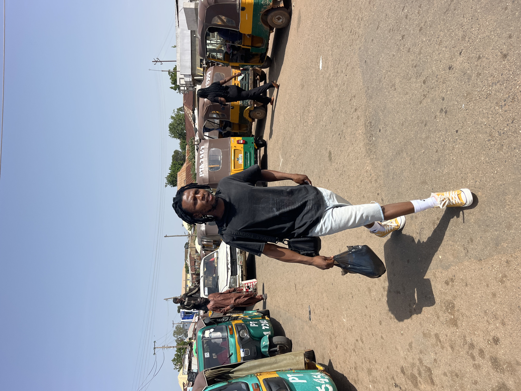 A young man with dreadlocks, wearing a black t-shirt and light blue shorts, stands on a paved road. He is holding a black plastic bag in his right hand and is facing the camera with a slight smile. His left leg is extended forward, and his right leg is bent at the knee, giving the impression he is in mid-stride or posing. He is wearing white socks with yellow sneakers.
The background shows a street scene with several auto-rickshaws, commonly known as "tuk-tuks," parked in a row. Some of these vehicles are green and yellow, while others appear to be yellow and brown. One tuk-tuk has "PTA 3967" and "LL 8557" visible on its license plate. Another person in a brown traditional outfit is walking away from the camera in the middle ground, between the auto-rickshaws. Further in the background, buildings and trees are visible under a clear, bright sky, suggesting a sunny day. Power lines and utility poles are also present. The overall setting appears to be an urban or semi-urban area, possibly a marketplace or a transportation hub. The lighting suggests it is daytime.