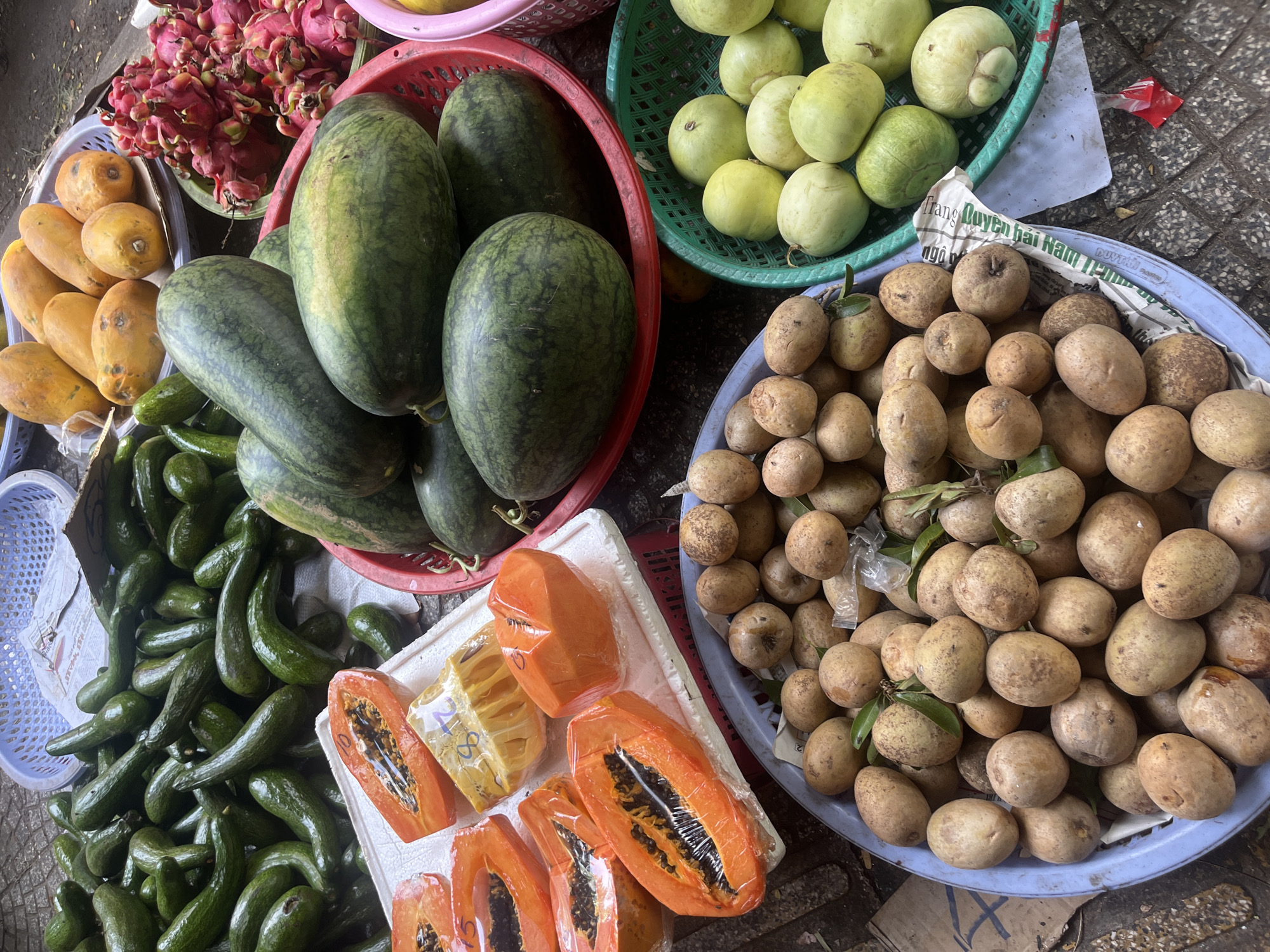 This image depicts a fruit stall at a market in Da Nang, Vietnam. The scene is a close-up, overhead view of various fruits arranged in baskets and on a white styrofoam base. In the foreground, a blue plastic basket is filled with numerous small, brown, oval-shaped fruits that appear to be sapodillas. To the right of the sapodillas, a green plastic basket contains several light green, round fruits, possibly guavas. Behind these, a red plastic basket is overflowing with several large watermelons. A pile of avocados is visible in the upper left corner, contained within a light blue plastic basket. In the center of the image, slices of bright orange papaya, some wrapped in plastic, are displayed on a white styrofoam base, alongside a piece of yellow fruit, likely jackfruit, also wrapped in plastic. Further back, a basket of yellow papayas and a basket of vibrant pink dragon fruits are visible. The ground appears to be paved with stone tiles. No people are visible in the frame.