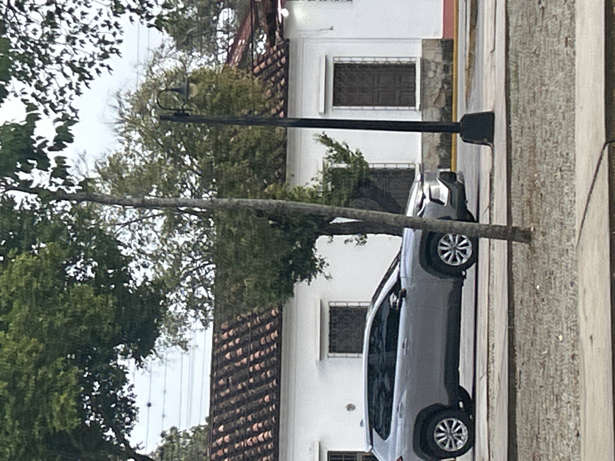 The image captures a street scene in Santa Ana, Venezuela. A silver SUV is parked on the right side of the frame, facing left. Behind the vehicle, a white building with a red tile roof is visible. The building has several windows with dark shutters. A black lamppost stands in front of the building. Lush green trees with dense foliage are scattered throughout the scene, with some branches extending over the building and the street. The ground in the foreground appears to be paved with concrete and covered with gravel. The sky is overcast, suggesting a cloudy day. There are no people visible in the image.