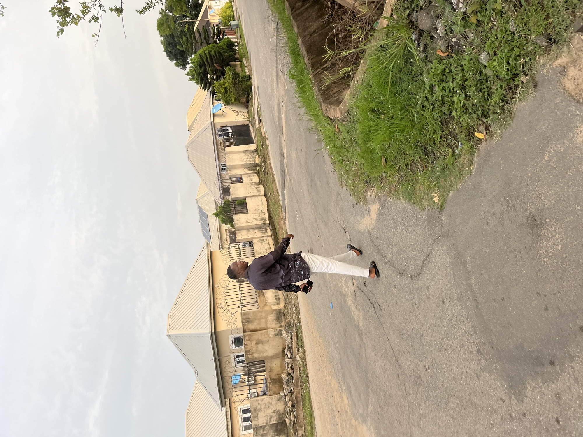 An adult male walks away from the camera on a paved road in Abuja, Nigeria. He is wearing a dark, patterned long-sleeved shirt, white pants, and black sandals. He is holding a black object in his right hand, possibly a phone. He is walking past several single-story houses with beige walls and light-colored, peaked roofs. The houses are bordered by fences with barbed wire. There is a drainage ditch with green grass and some debris on the right side of the road. The sky is overcast.