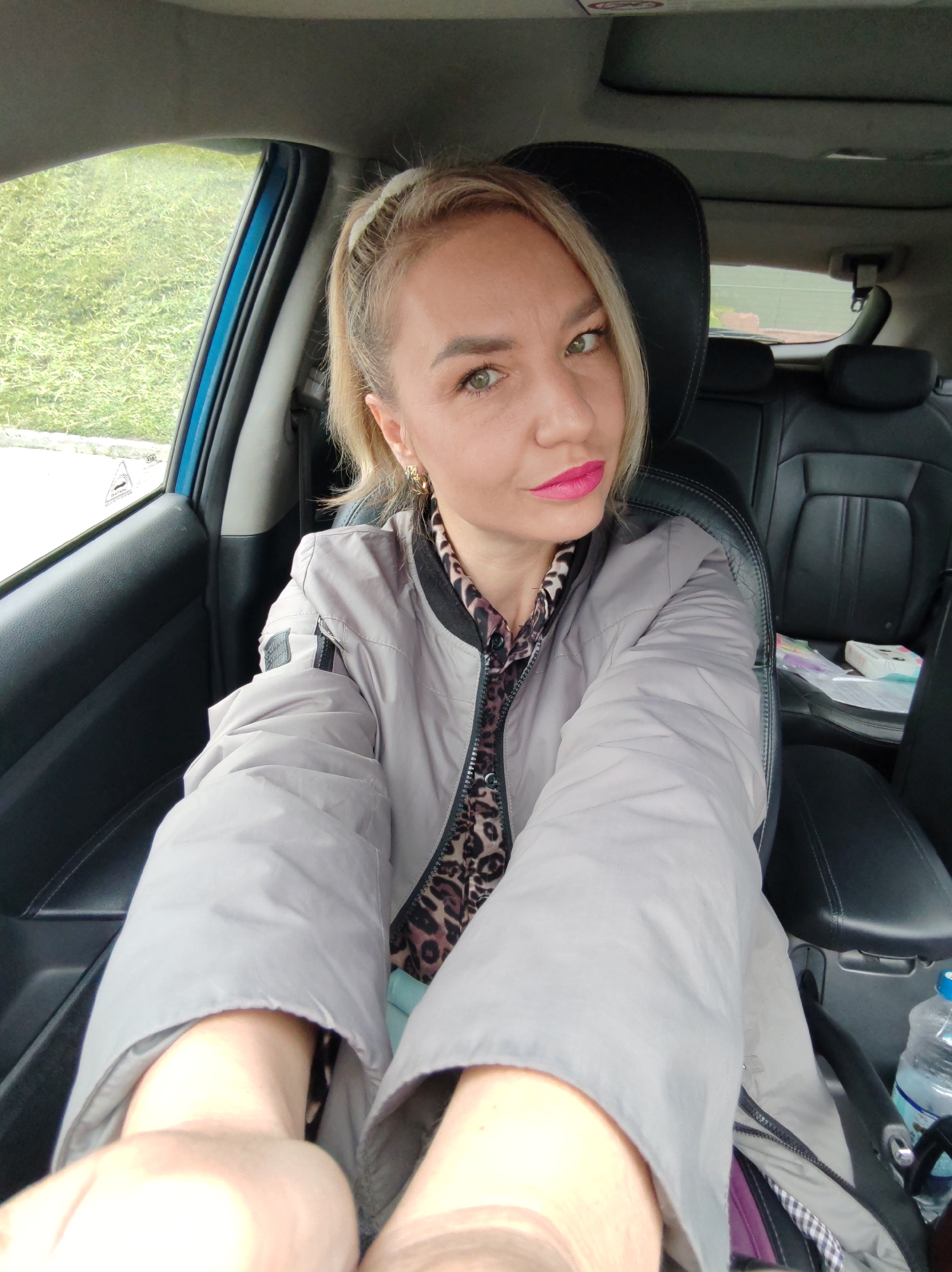 A woman is sitting in the driver's seat of a car, taking a selfie. She has blonde hair pulled back in a ponytail and is wearing a light gray jacket over a leopard-print top. Her lips are painted bright pink. The car's interior is visible, with black leather seats and a blue door frame. Outside the car, a green grassy area is visible through the window. A water bottle is on the passenger seat.