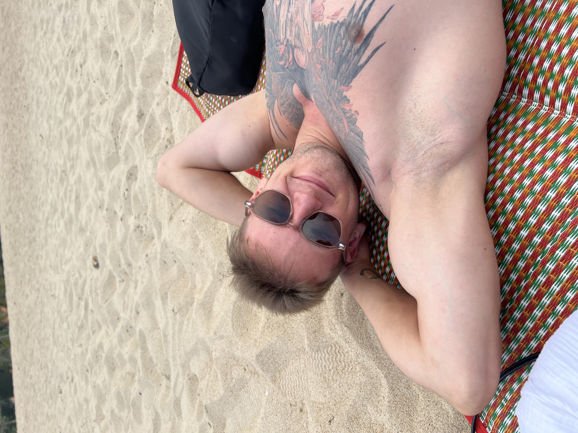 A man is depicted lying on his back on a sandy beach, seen from a high-angle perspective. He is positioned diagonally across the frame, with his head towards the bottom center and his body extending towards the upper right. His hands are clasped behind his head, and he is wearing reflective sunglasses. He looks directly at the camera with a relaxed smile.
A large, detailed tattoo, appearing to be a dark-colored bird or winged creature, covers a significant portion of his chest and left shoulder. He rests on a colorful, woven mat with a repeating pattern of red, green, and white stripes. To his upper left, a black bag is visible on the mat. The surrounding area is light-colored sand, showing subtle patterns and textures from wind or water. The bright lighting suggests it is daytime, indicating sunny weather. The scene conveys a leisurely moment, consistent with a beach setting like Nha Trang Ward, Vietnam. There is no visible text in the image.
