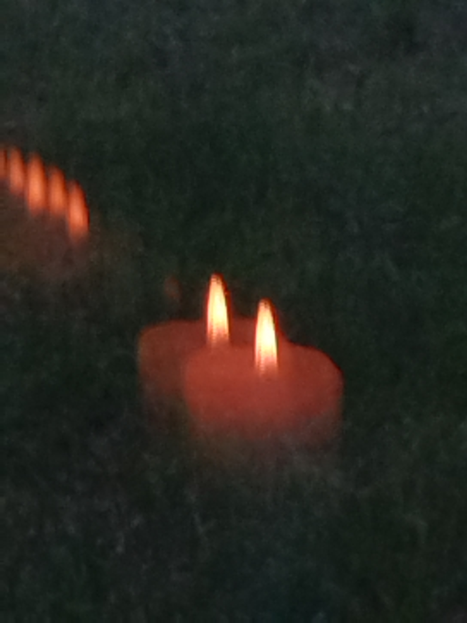 The image depicts a row of flickering candles, with two in focus in the foreground and others blurred in the background, arranged on a dark, grassy surface. The candles are lit, emitting a warm orange glow. The overall impression is dim, likely taken at dusk or night. The scene is devoid of any human presence or identifiable urban structures.