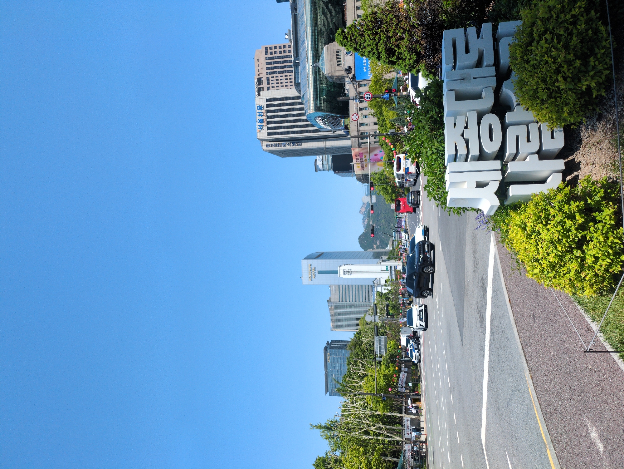 The image shows a street view in Seoul, South Korea, captured on a sunny day. The sky is a clear blue. The camera is positioned alongside a road and a sidewalk, with a large white sculpture of Korean characters and landscaping visible to the right.
The road is grey and lined with white markings. Multiple vehicles, including cars and a police van, are seen traveling on the road, which extends into the distance. Buildings of varying heights and designs are visible on both sides of the road, with some appearing to be modern skyscrapers. A building called “Koreana Hotel” can be seen. Trees are interspersed between the buildings. Pedestrians appear along the sidewalk. A speed limit sign is visible on the street, marked "50". In the background, distant mountain peaks are visible.