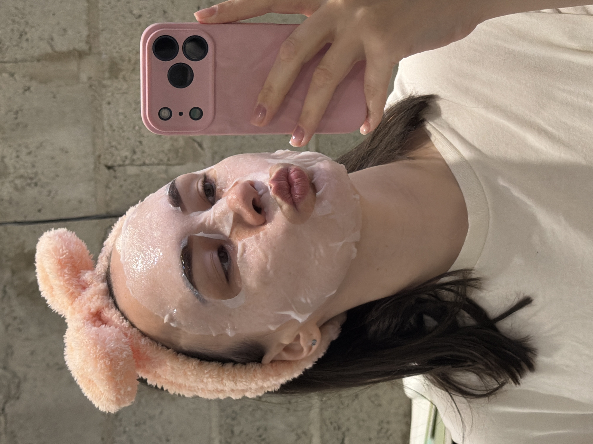 A person is holding a pink smartphone to take a selfie. They have a pink headband with bunny ears and a sheet face mask on. The person is wearing a white t-shirt. The background appears to be a gray wall. There is no information to suggest this image was taken in Chaikovsky, Russia.