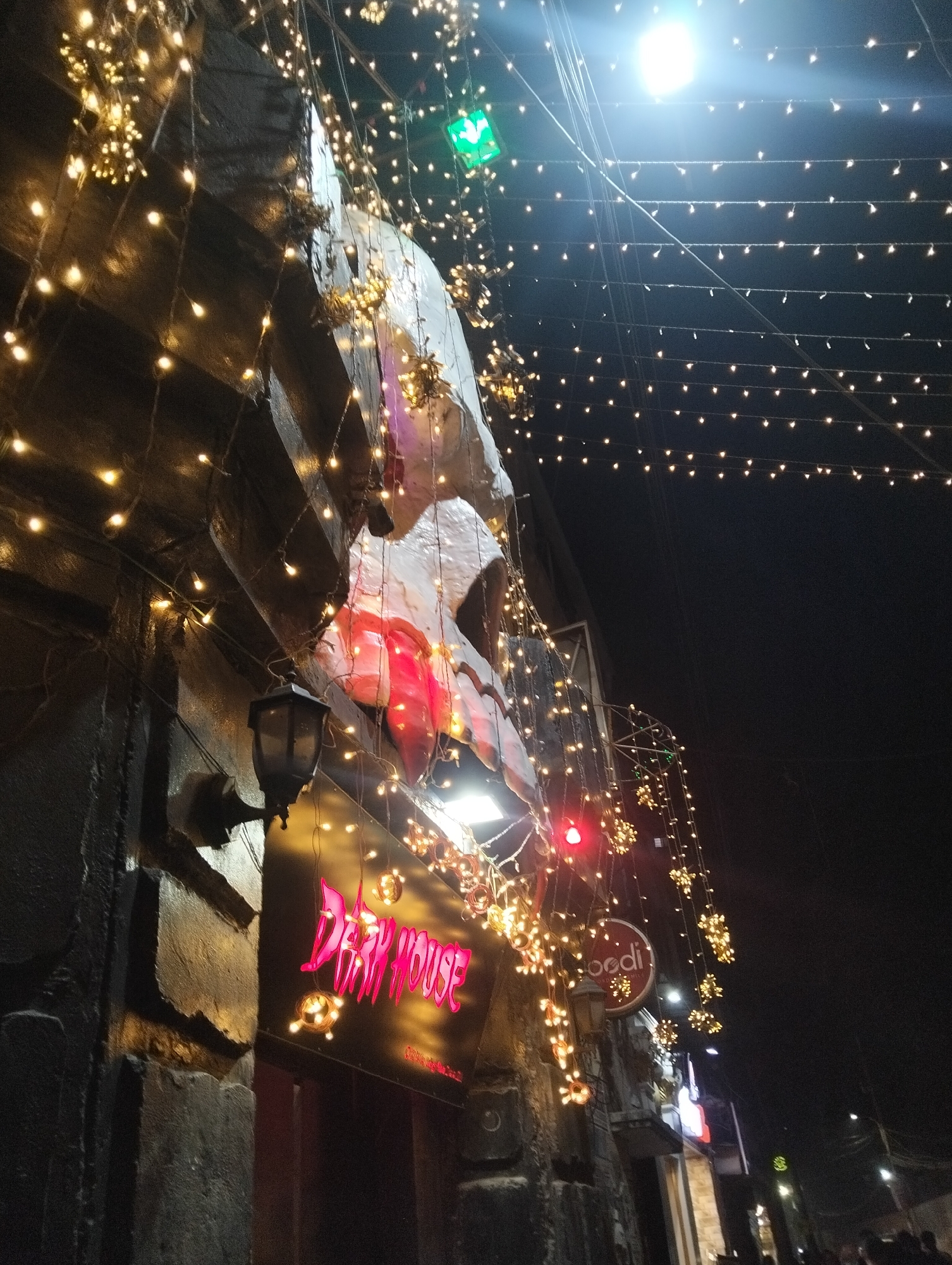 A nighttime image depicts the "Dark House" establishment in Dhaka, Bangladesh, decorated with string lights. The building's exterior is stone or masonry. A lantern-style light fixture is mounted on the wall near the entrance. A sign for "Dark House" is illuminated in pink neon. Above the sign is a lighter-colored, possibly sculptural element. Additional illuminated signage is visible further down the street. People are gathered in the distance. Overhead, a network of string lights is suspended, stretching across the frame.